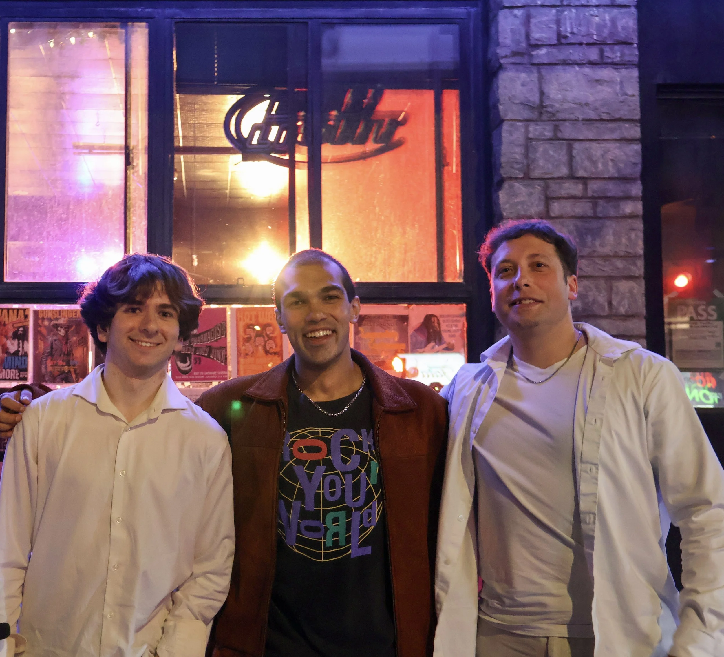Three men smiling and posing for a photo outside a building at night. The background features a brick wall, large windows revealing colorful neon and indoor lighting, and posters.