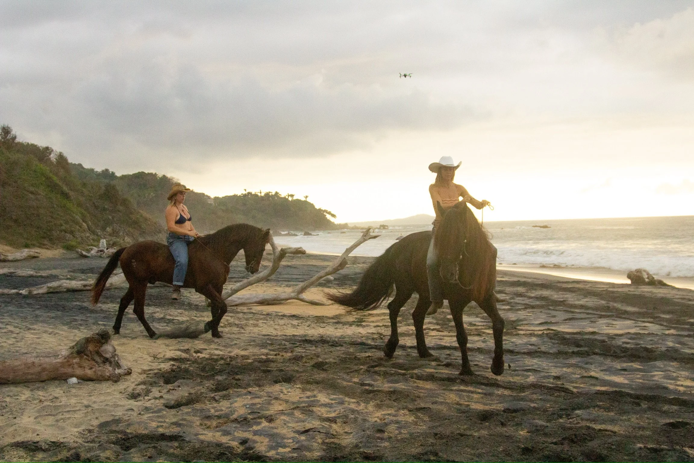 Two women riding horses along a beach at sunset, with a cloudy sky and distant hills in the background.