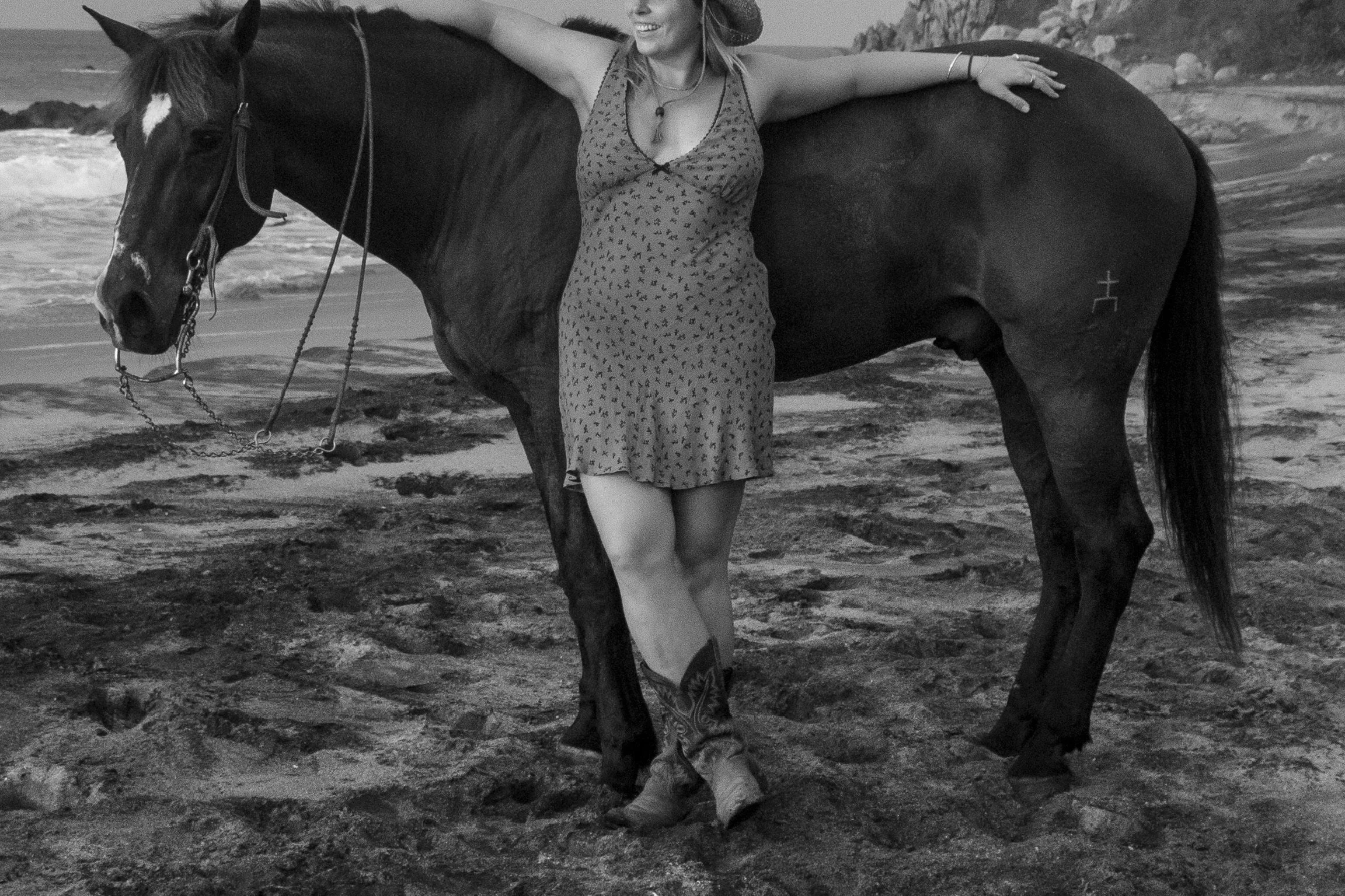 A woman smiling and wearing a hat, standing on a beach with her arm around a horse, with rocky cliffs in the background.