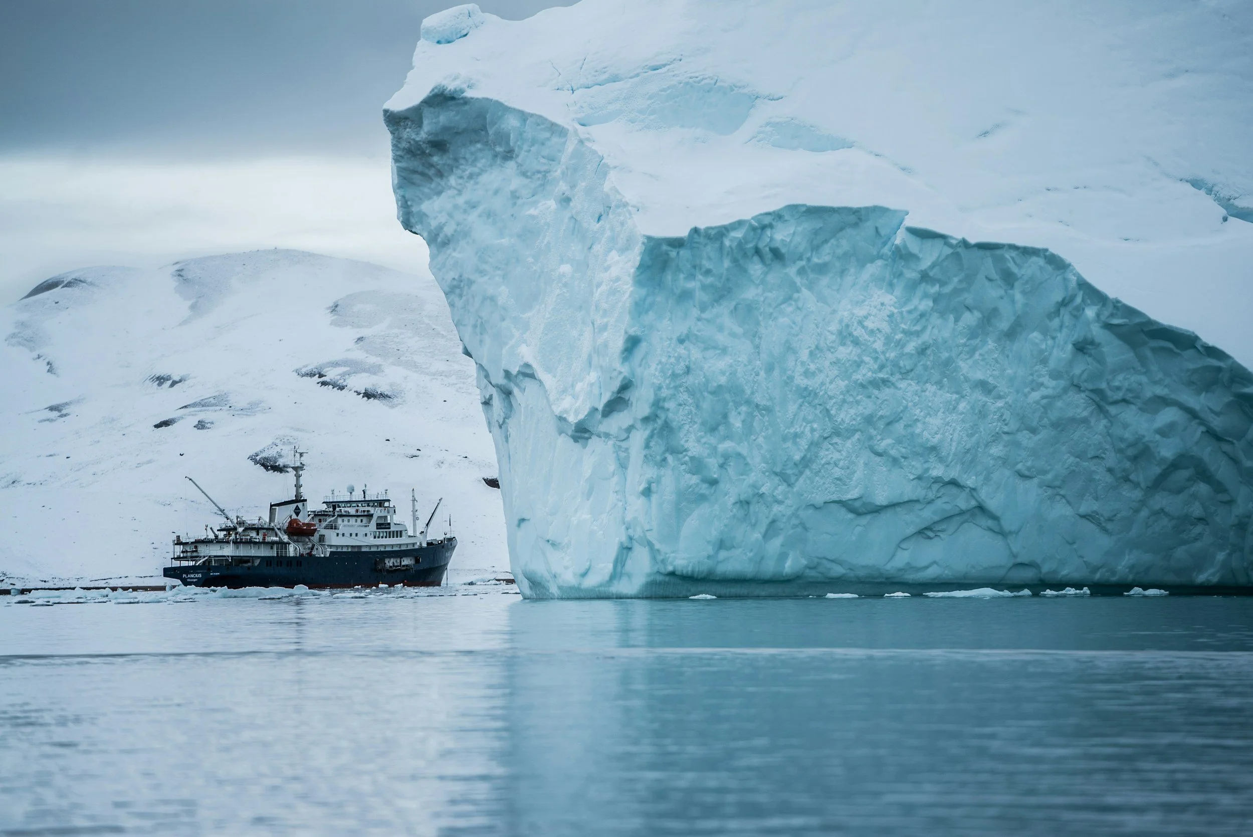 A large iceberg floating in icy waters with a ship in the background, surrounded by snow-covered landscape and overcast sky.