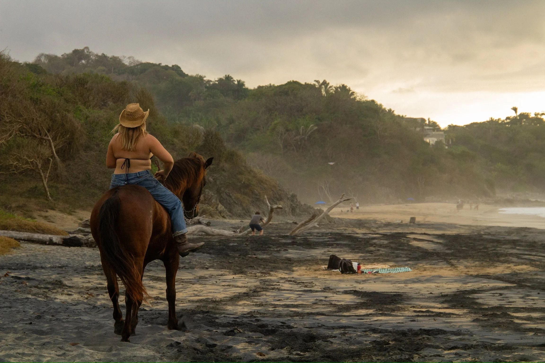 A person with blonde hair wearing a straw hat, a bikini top, and jeans riding a brown horse along a beach at sunset, with a hill covered in greenery in the background.