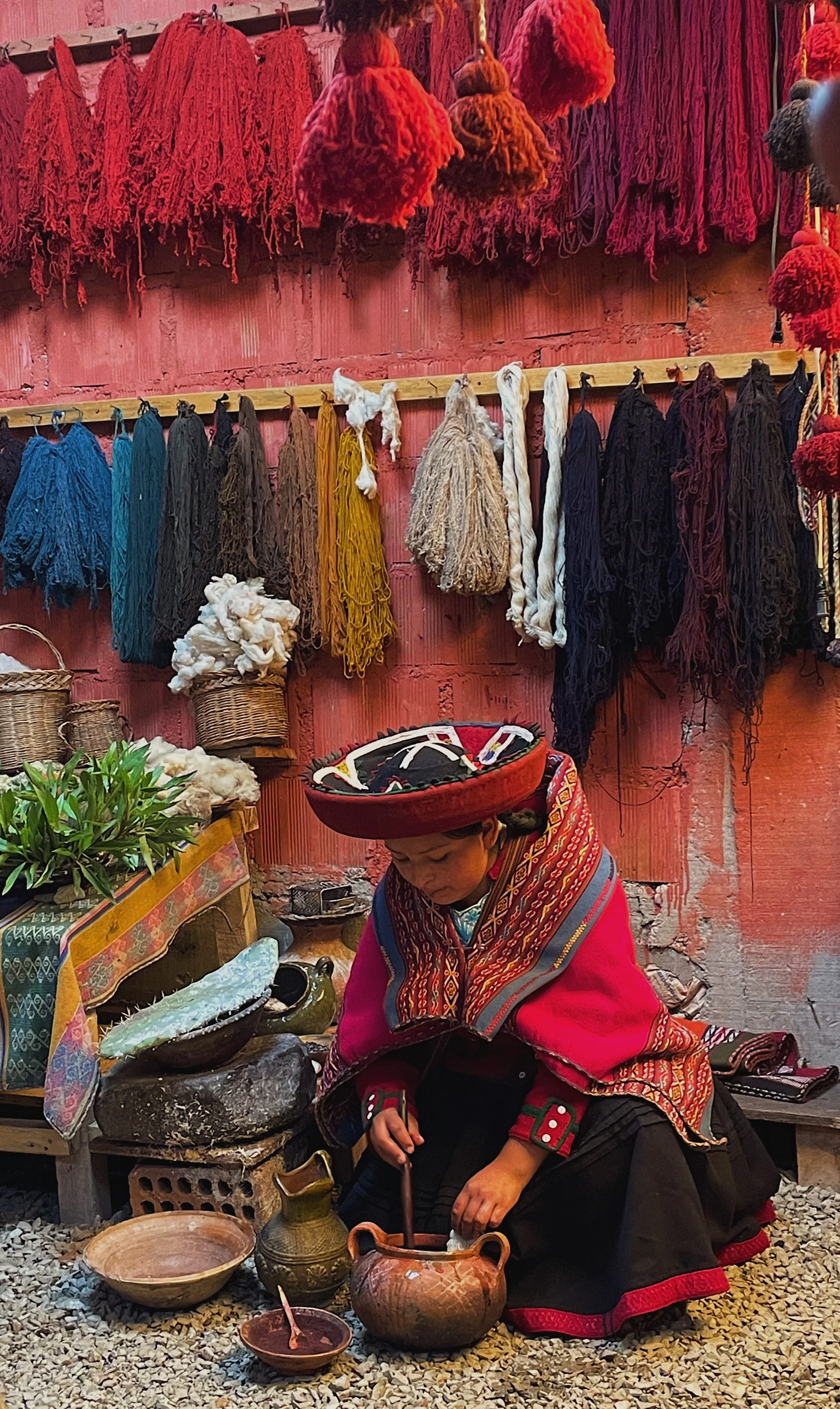 A woman dressed in traditional indigenous clothing, sitting on the ground and working with pottery. Behind her, shelves display colorful yarn and textiles, with baskets and crafts around her.
