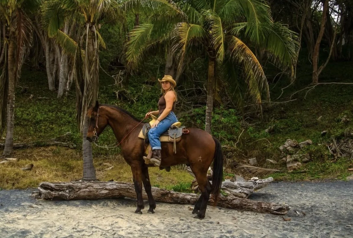 Woman riding a horse on a sandy beach with palm trees and lush greenery in the background.