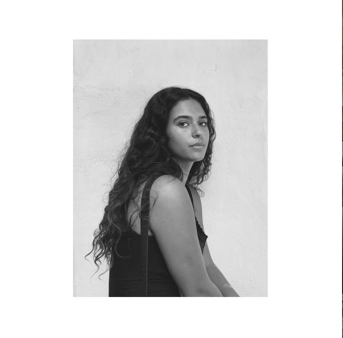 Black and white portrait of a young woman with long, curly hair, sitting against a plain light-colored wall, wearing a sleeveless top, looking at the camera with a neutral expression.