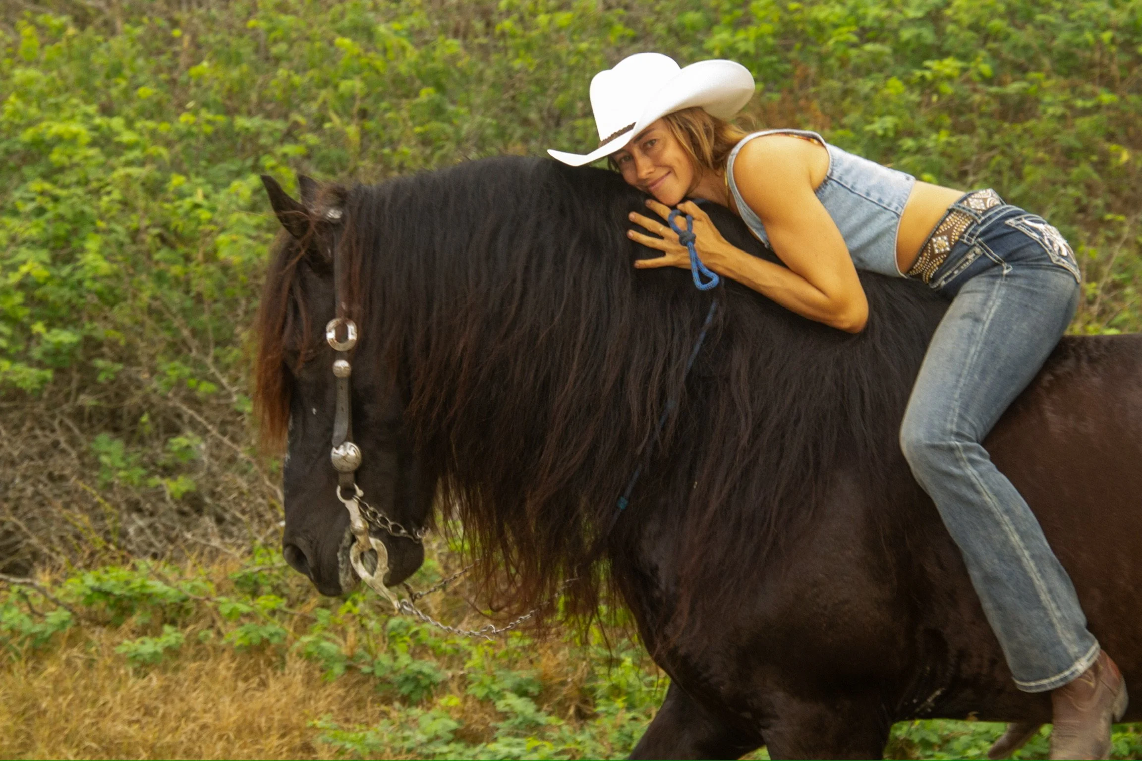A woman with a white cowboy hat hugging a black horse in a forested area.