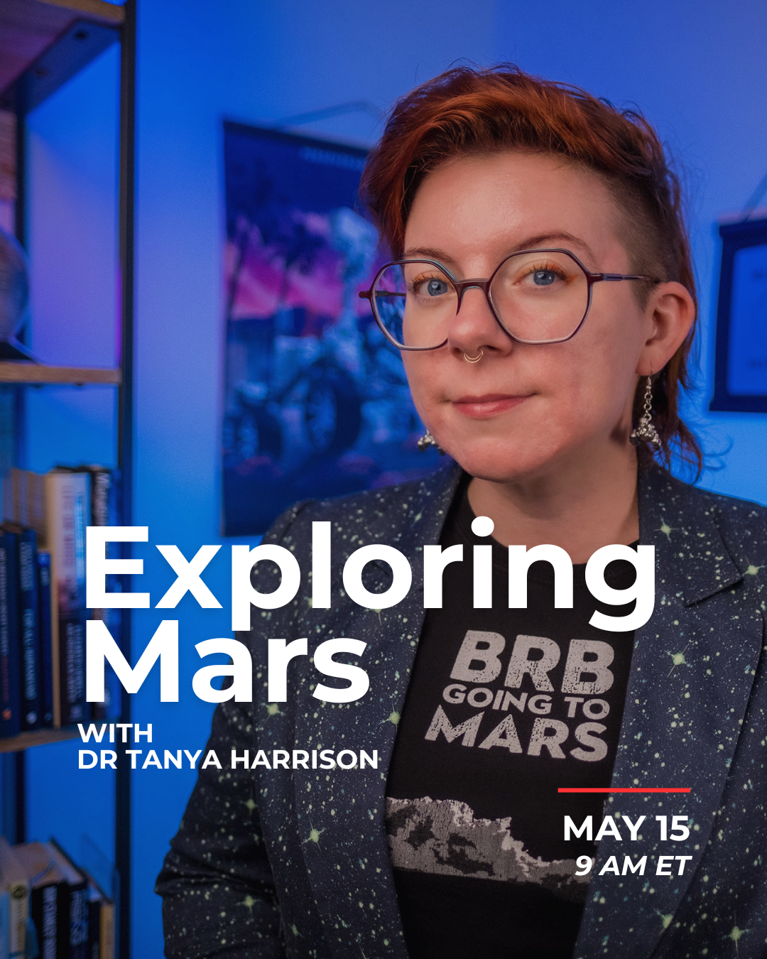 A woman with short, partially shaved reddish hair, wearing glasses with round frames, earrings, a septum piercing, a black t-shirt with an illustration and text, and a blazer with a galaxy pattern. She is in a room with blue lighting and a bookshelf in the background.