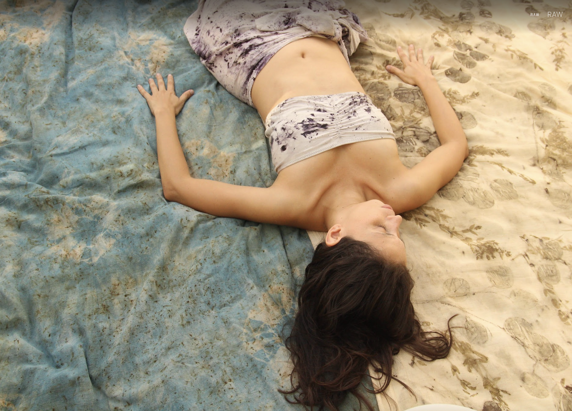 A woman lying on her back on a bed with a patterned blanket, her eyes closed and arms resting at her sides.
