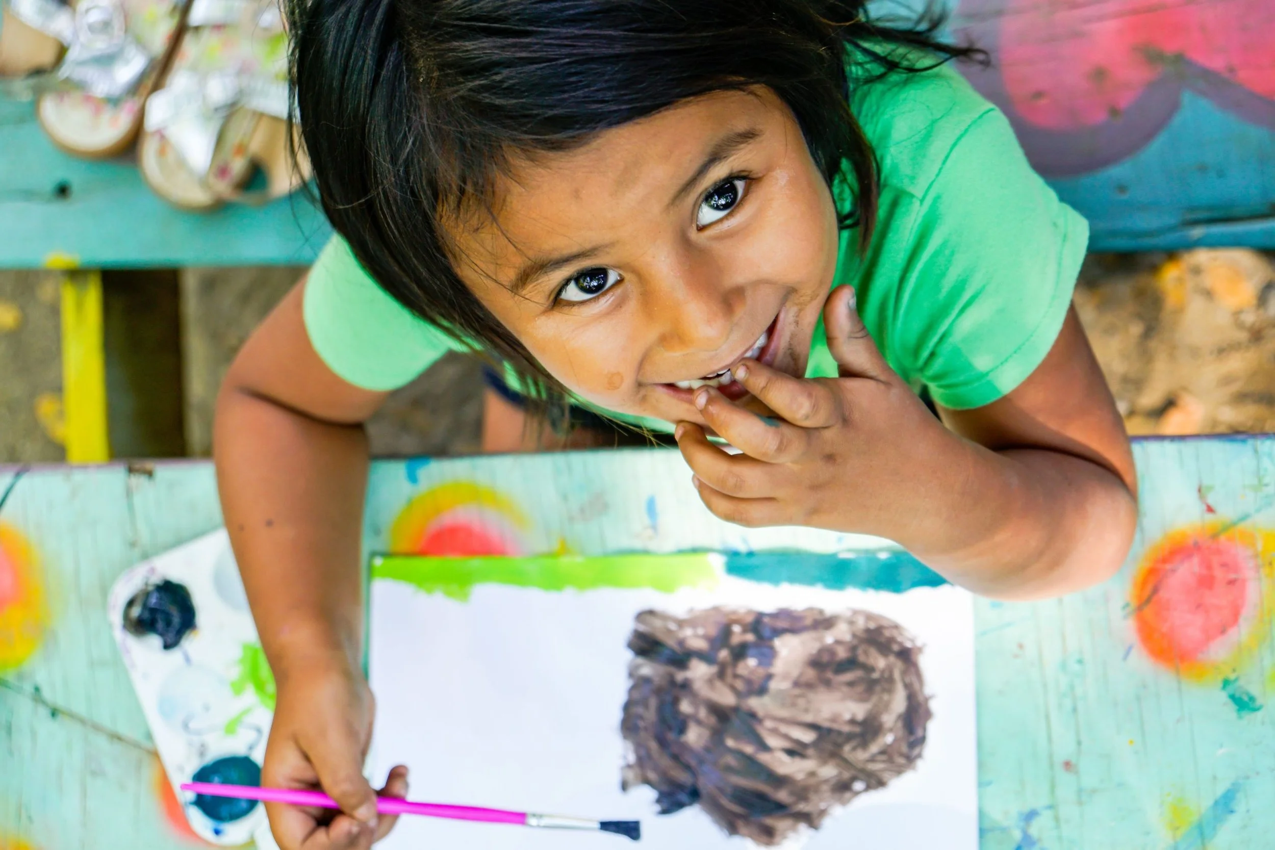 Young boy with dark hair and brown skin painting at an outdoor table, wearing a green shirt, with colorful paint and brushes on the table.