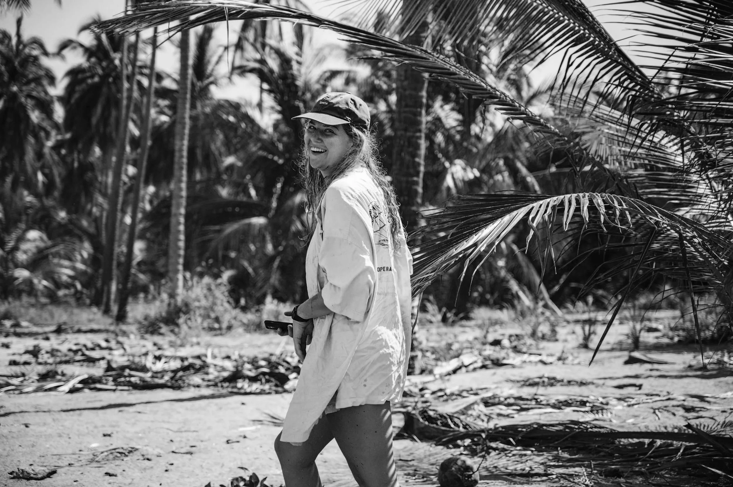 A woman smiling and holding a phone stands in a tropical setting with palm trees.