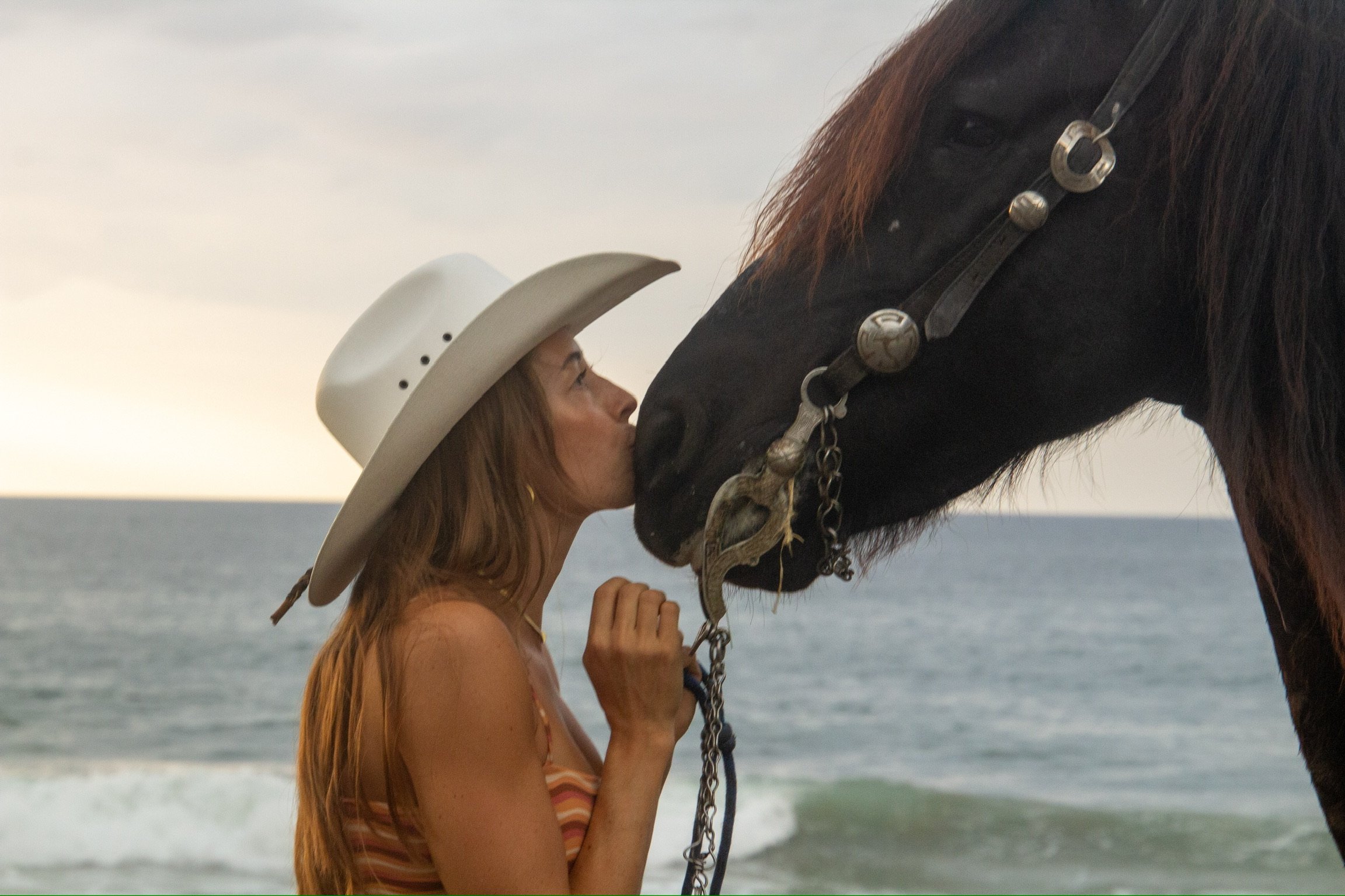 A woman with long reddish hair wearing a cowboy hat, standing on the beach, kissing the nose of a large black horse that has a bridle and chain, with the ocean and sky in the background.
