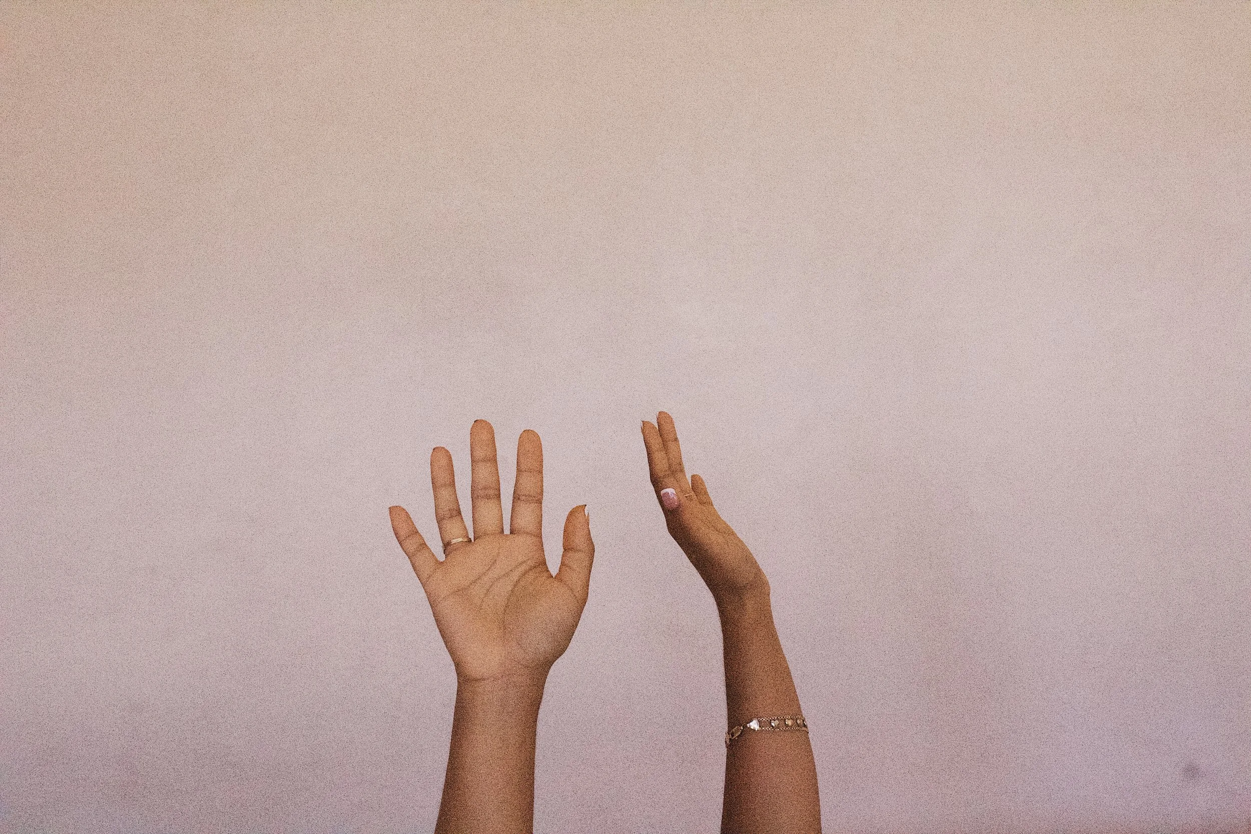 Two hands reaching up towards a pinkish wall, with one hand wearing rings and a bracelet.