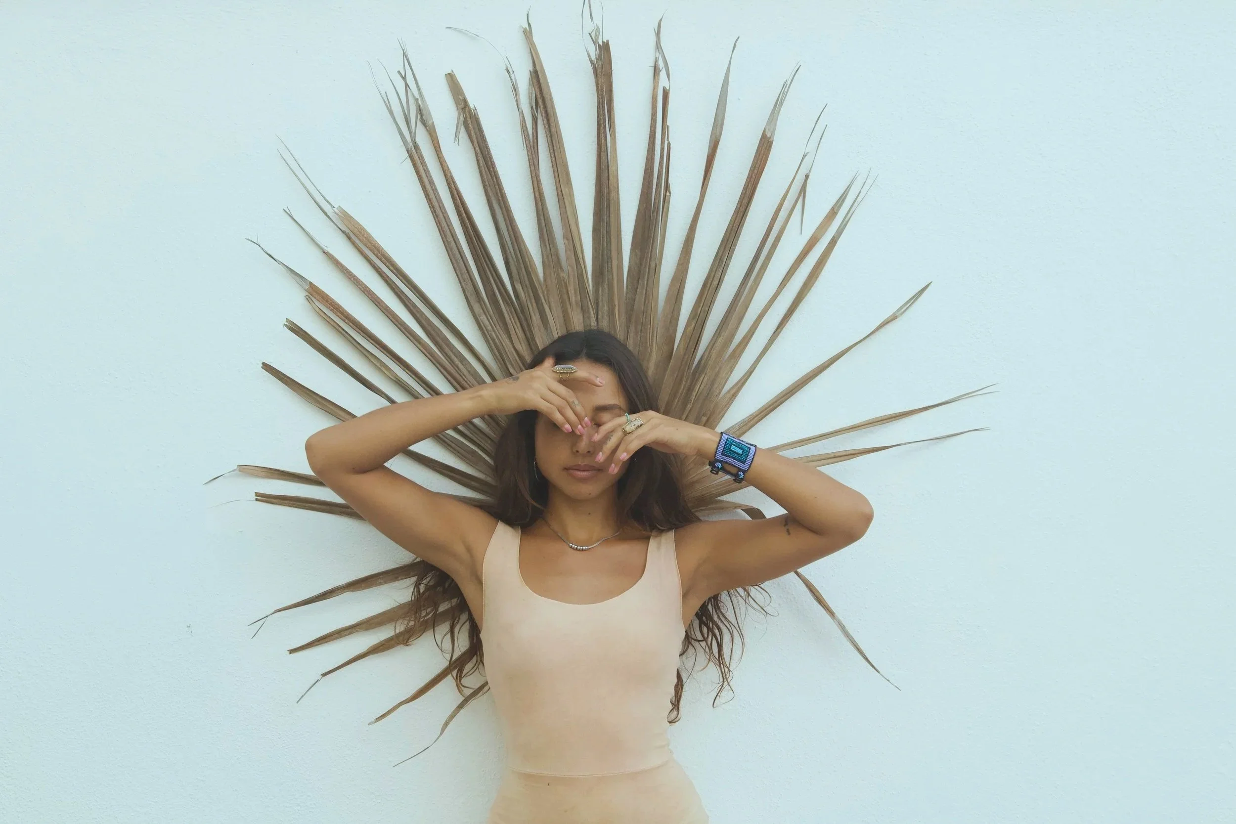 A woman with long brown hair lying on a white wall with dried palm leaves arranged in a radiating pattern behind her. She is wearing a beige tank top, multiple rings, a bracelet, and a necklace, with her hands covering her eyes.