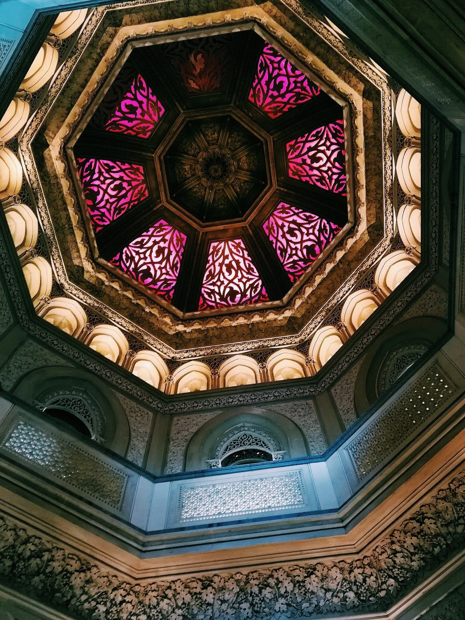 The image shows an ornate, octagonal ceiling with intricate patterns illuminated in pink and purple lights, surrounded by decorative architectural details and balconies.