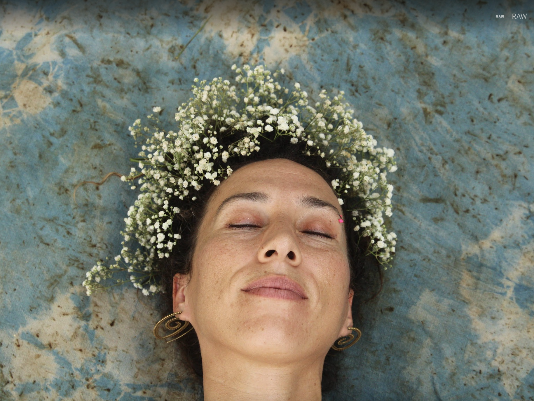 A woman with dark hair lying down with her eyes closed, wearing a floral crown made of small white flowers, on a blue textured surface.