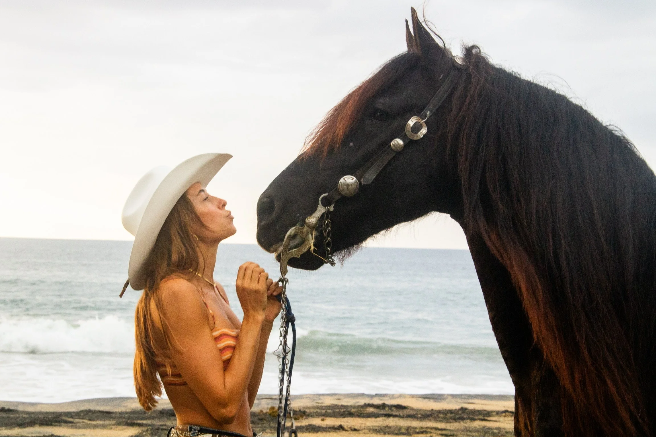 A woman with long reddish hair wearing a white cowboy hat, standing on a beach, facing a large black horse with a brown mane and wearing a bridle, as waves crash in the background.