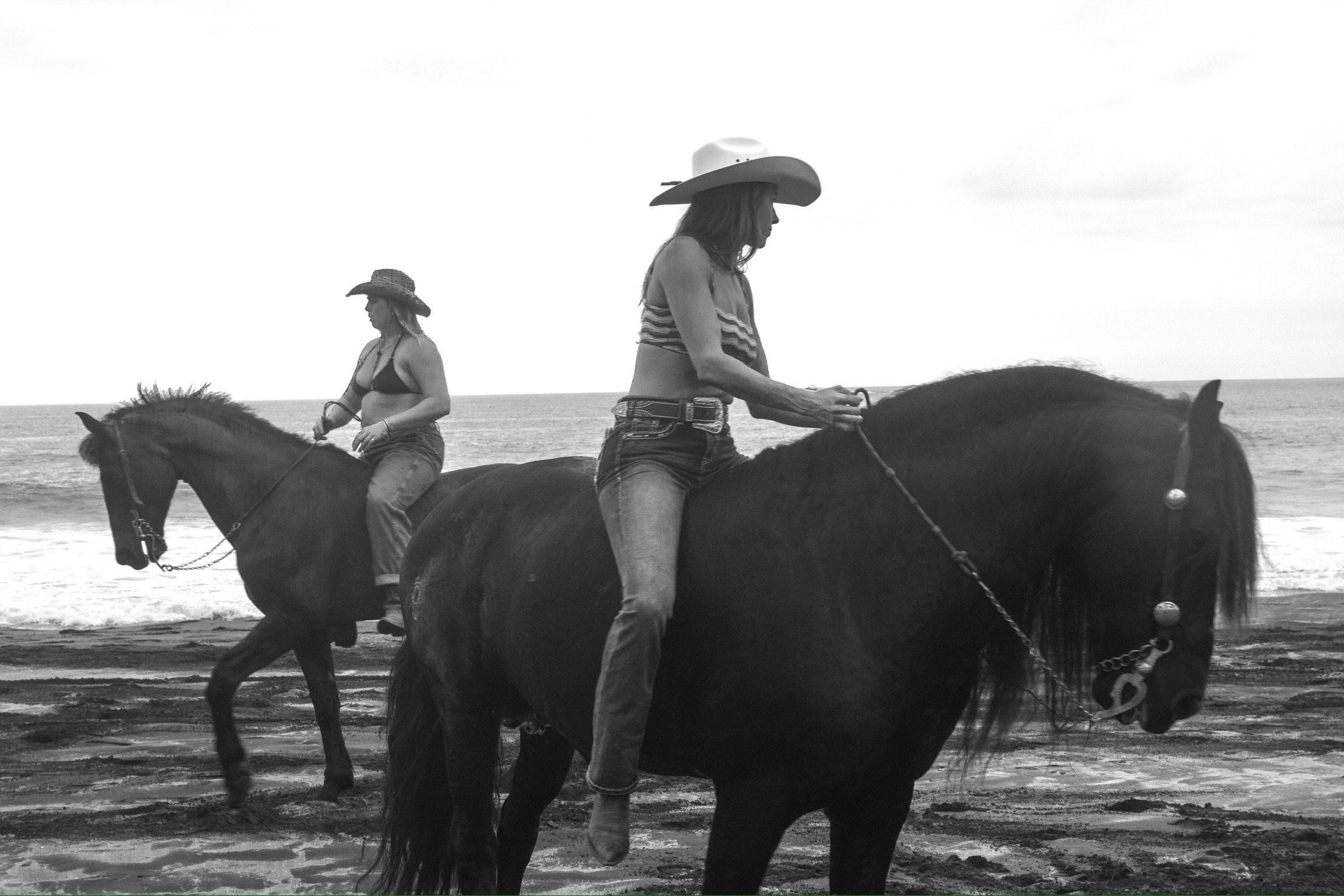 Two women riding horses along a beach, wearing cowboy hats and casual clothing, in black and white photo.