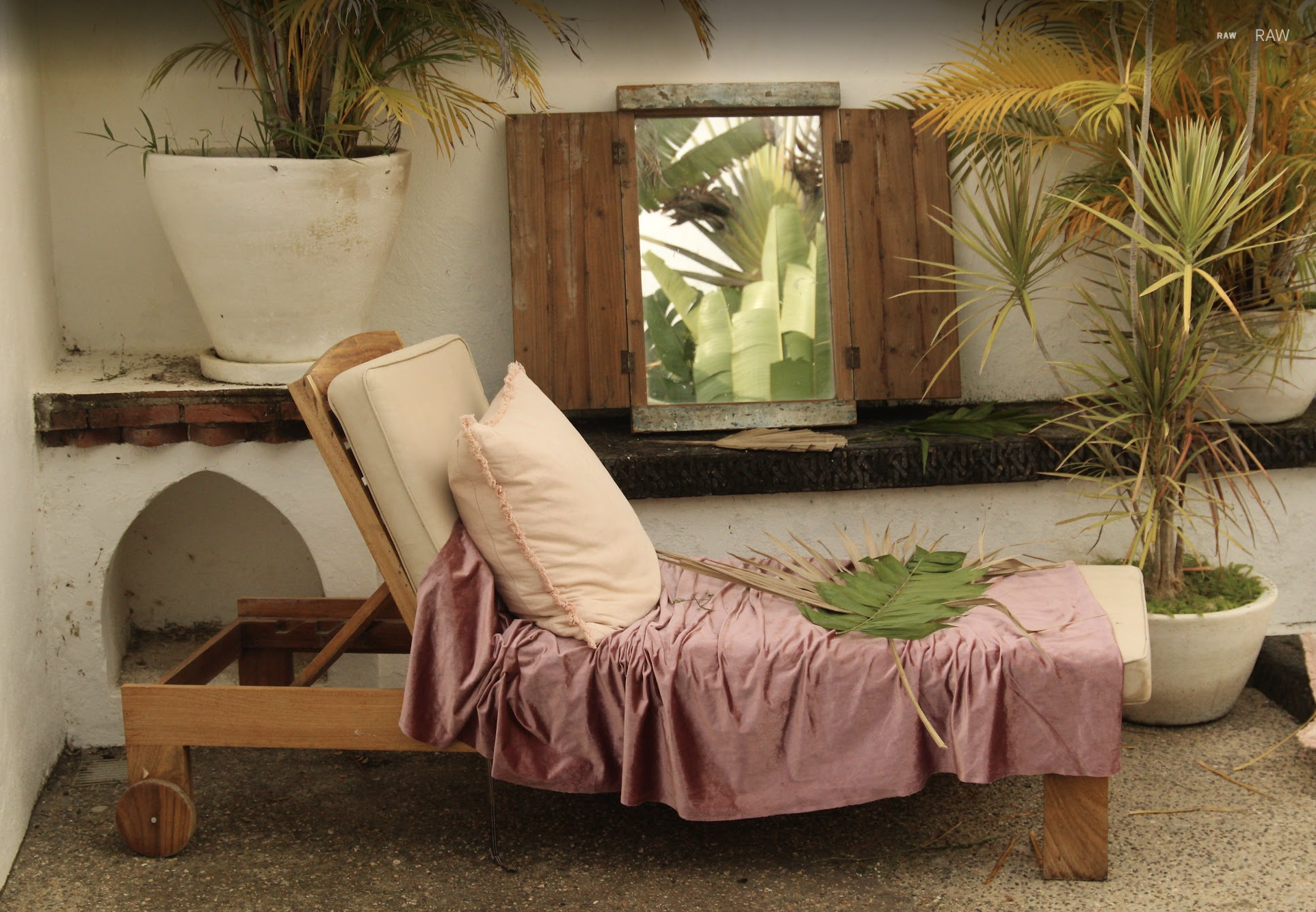 A rustic outdoor seating area with a wooden platform, pink fabric, and plush pillows. Large potted plants and a mirror with wooden shutters decoratively placed behind it add to the cozy, tropical ambiance.