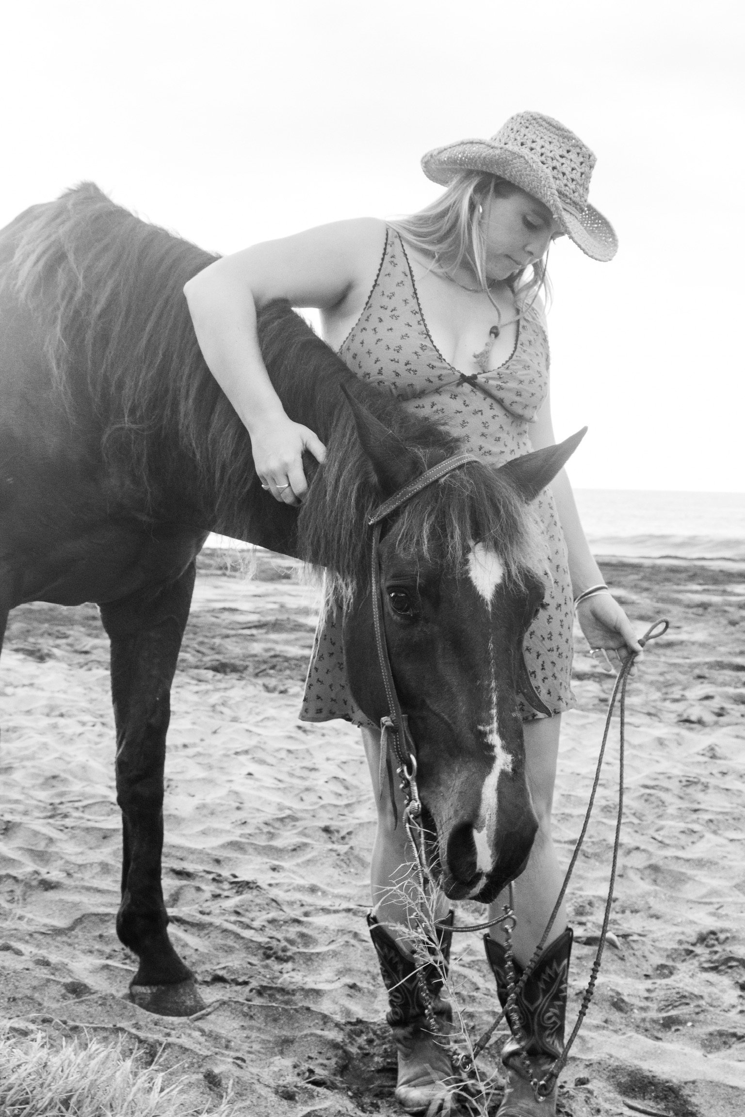 A woman wearing a straw hat, a patterned dress, and cowboy boots is standing on the beach, holding the bridle of a horse, which has a distinctive white blaze on its face.