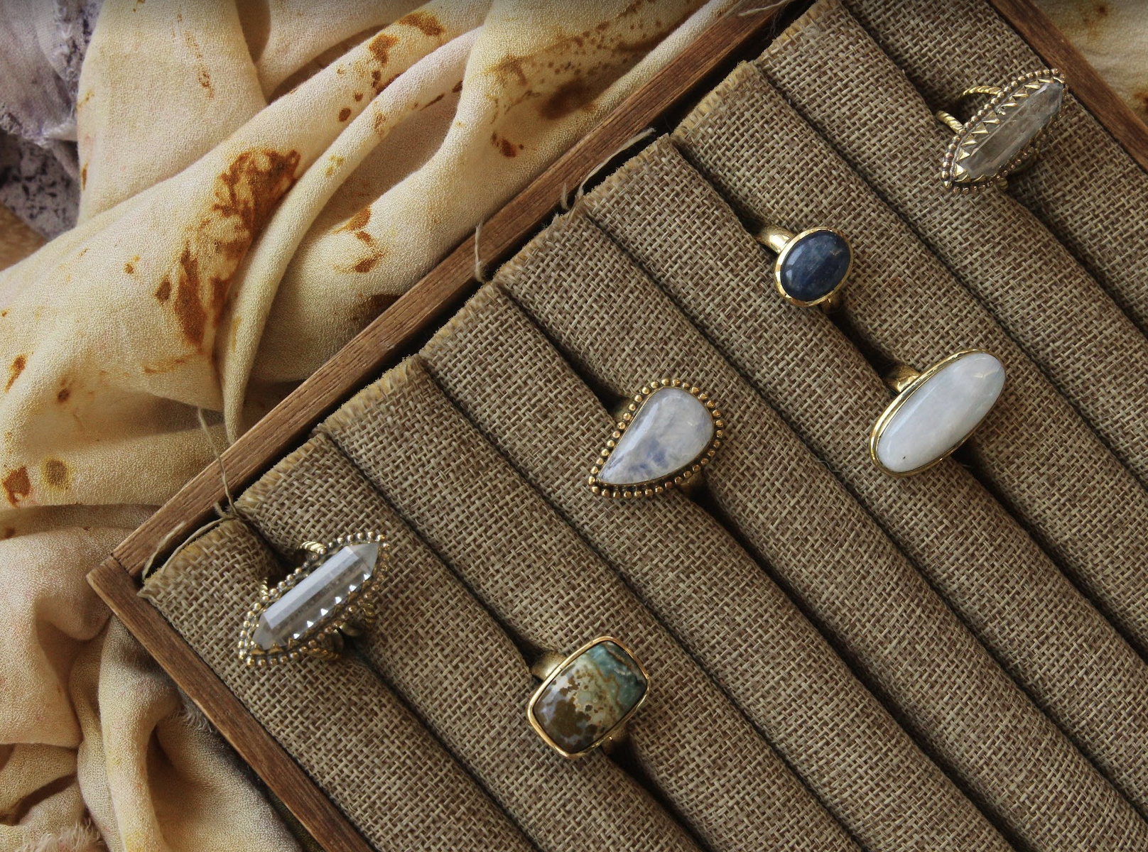 A display of various rings with large stones on a textured fabric surface, with floral patterned fabric in the background.