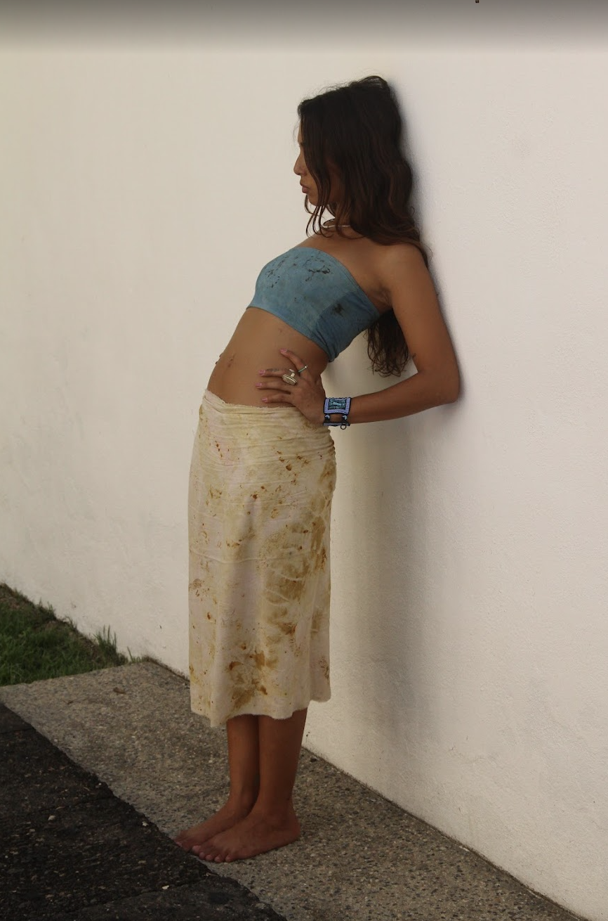 A woman with long wavy hair standing barefoot against a white wall, wearing a blue strapless top and a long beige skirt stained with dirt, with her hands on her hips and head tilted downward.