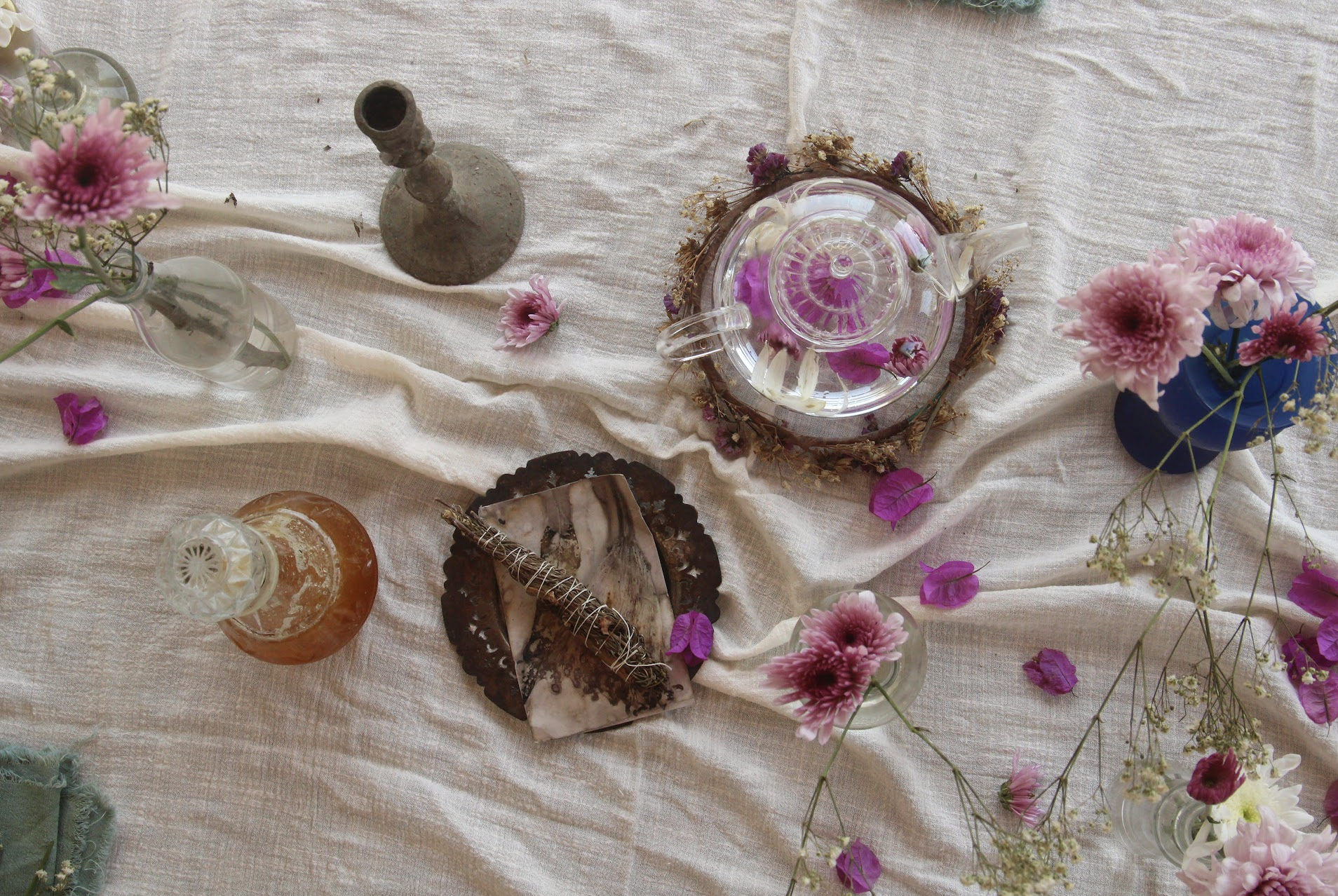 A top-down view of a decorative table setting featuring a clear glass teapot with pink flowers inside, surrounded by pink flowers, dried herbs, and assorted candles on a white textured tablecloth.
