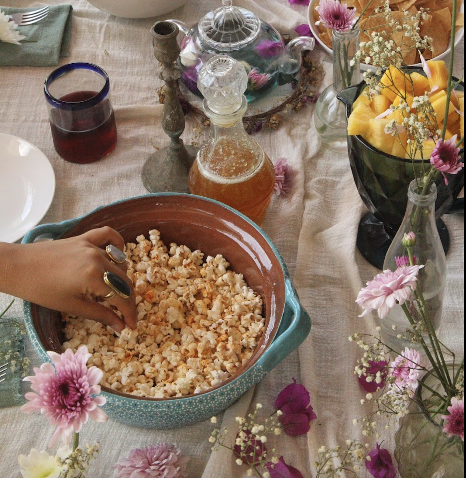 A table set with a large bowl of popcorn, floral arrangements in vases, a glass teapot, a growler, a glass of red wine, and plates and utensils in the background.