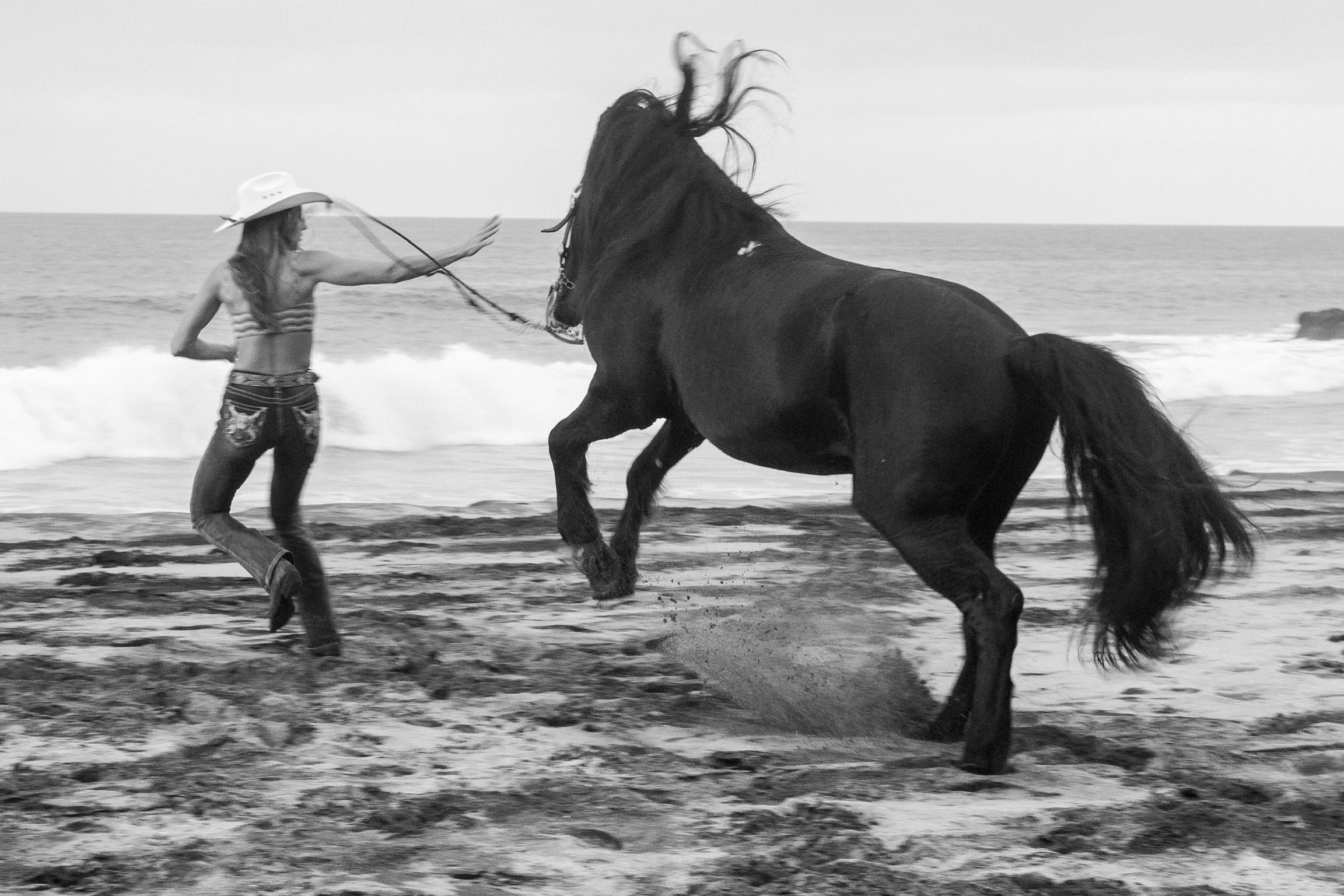 Black and white photo of a woman on the beach wearing a wide-brimmed hat and a striped swimsuit, reaching out towards a large black horse that is running or rearing.