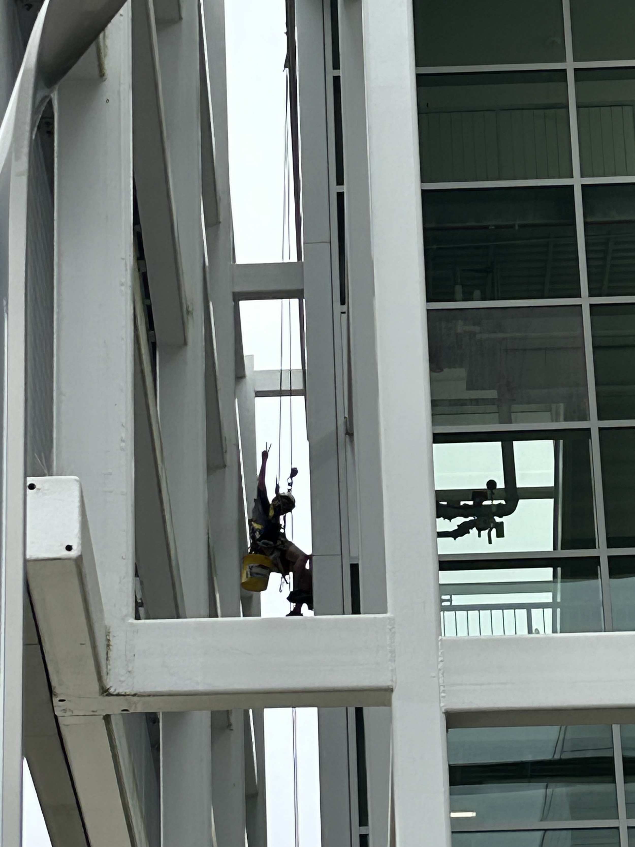 Professional window cleaner hanging from a harness off the side of a building, putting up a peace sign