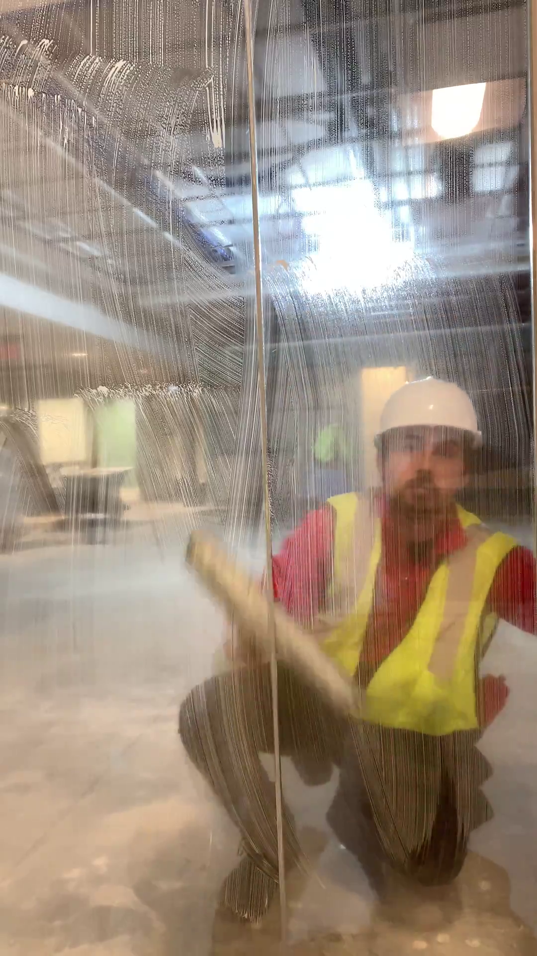 Professional window cleaner in a red uniform wearing a safety vest and helmet, behind a soapy pain of glass in a construction site.