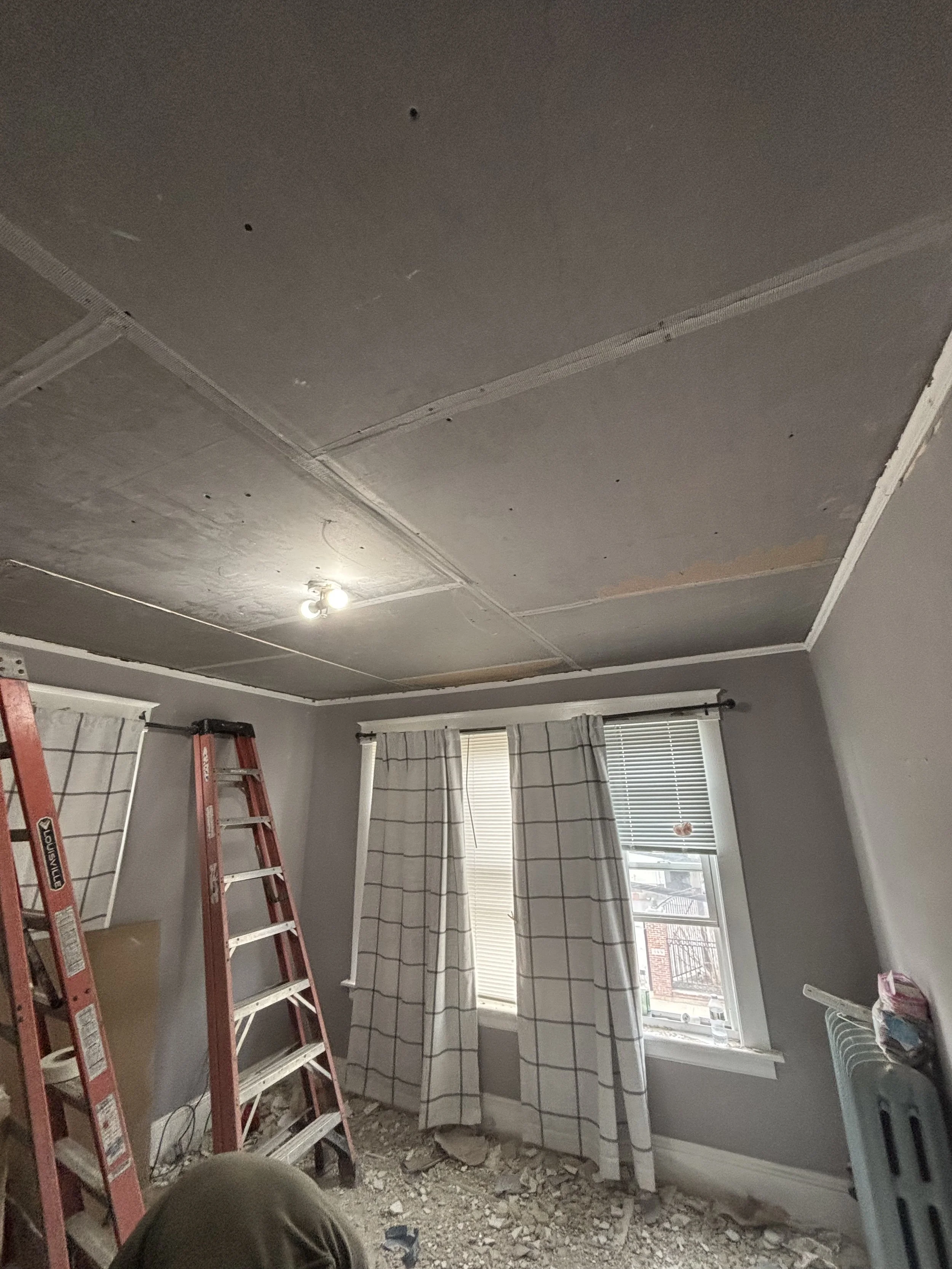 Room under renovation with a partially removed ceiling, a red ladder, and a window with curtains. Construction debris and a ladder are visible, and the ceiling has visible seams and holes.