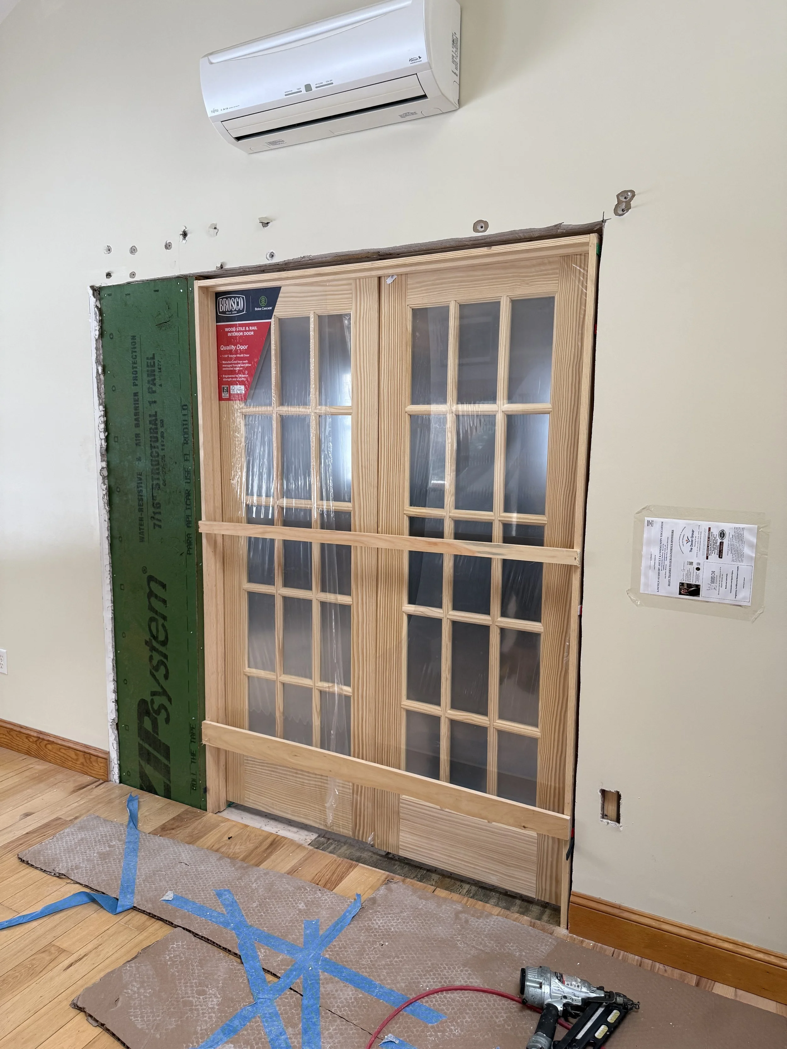 A wooden door with multiple glass panels is being installed in a wall under construction. A green insulation board and some tools, including a power drill and protective padding, are on the floor.