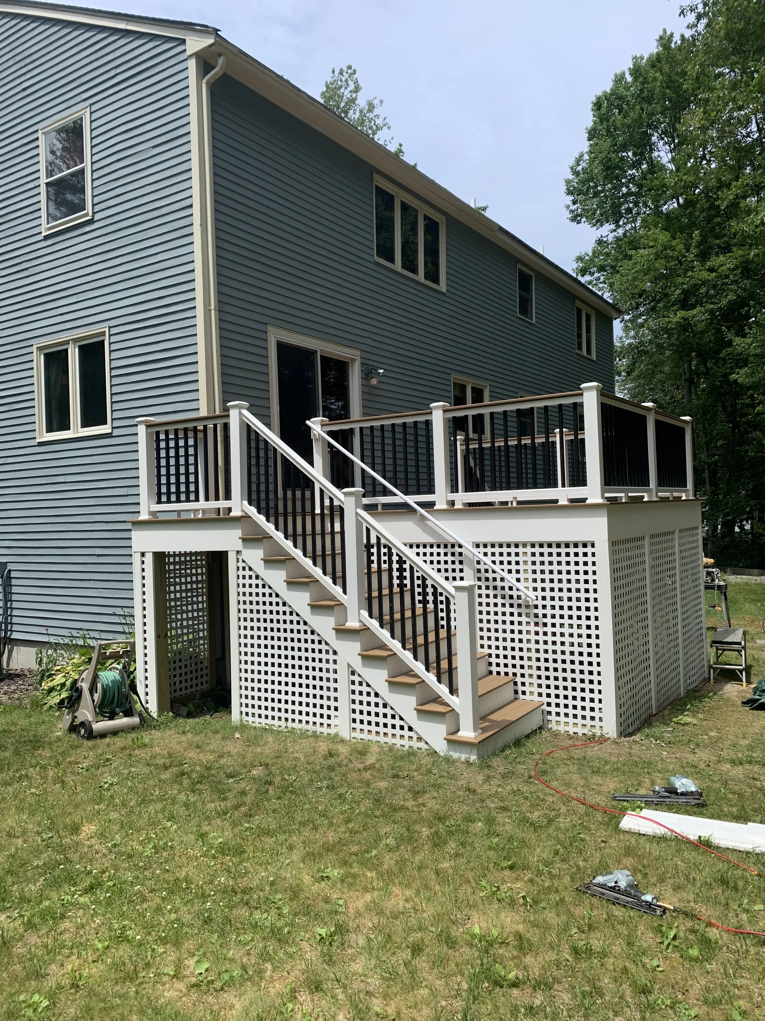 A two-story house with blue siding and white trim. There is a wooden deck with stairs leading to the backyard. The yard has some grass and tools scattered around, and trees are in the background.