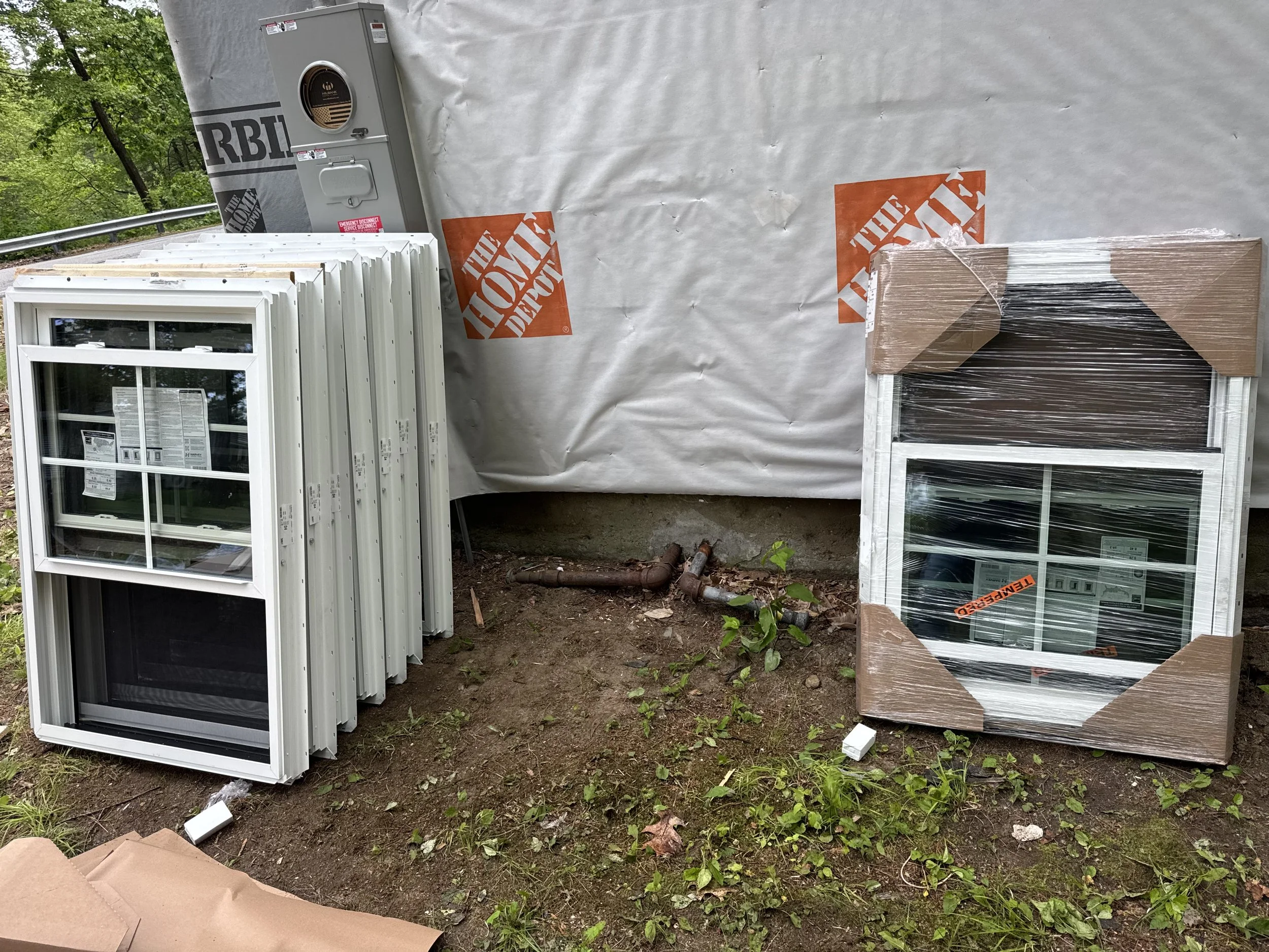 Bundled window frames, a packaged window, and installation materials outside near a house under construction, surrounded by trees and a metal guardrail.