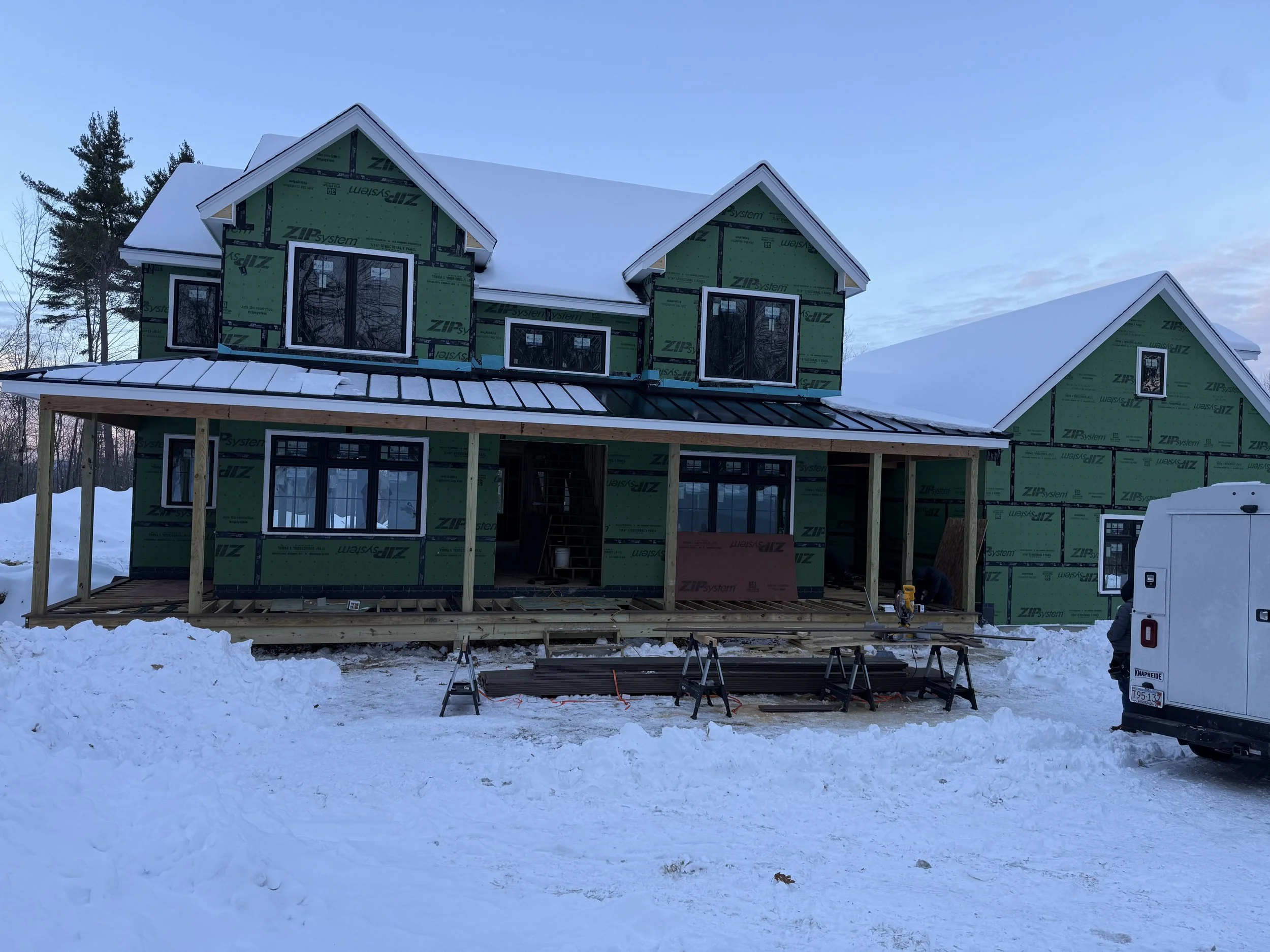 A house under construction with green sheathing and black window frames, situated in a snowy landscape. The house has multiple gabled roofs, and part of a deck is being built in front. Construction tools and materials are visible around the site.