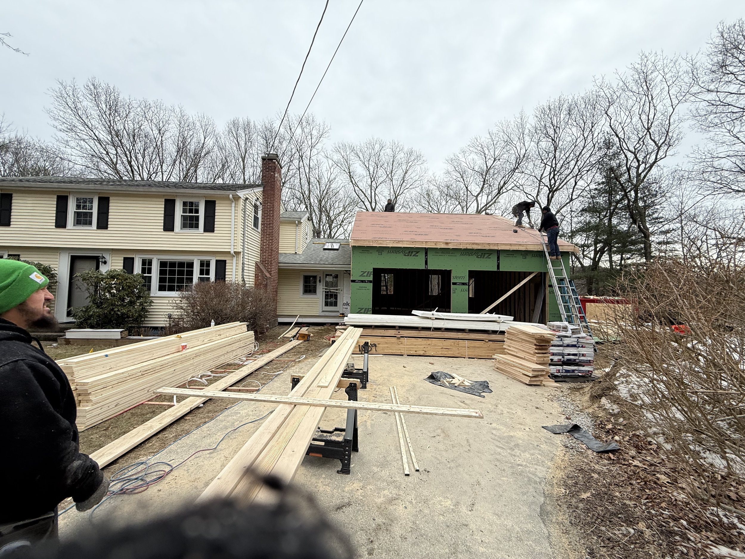 Construction workers building a roof on a residential house, with lumber and tools on the ground, in a suburban neighborhood with trees in the background.