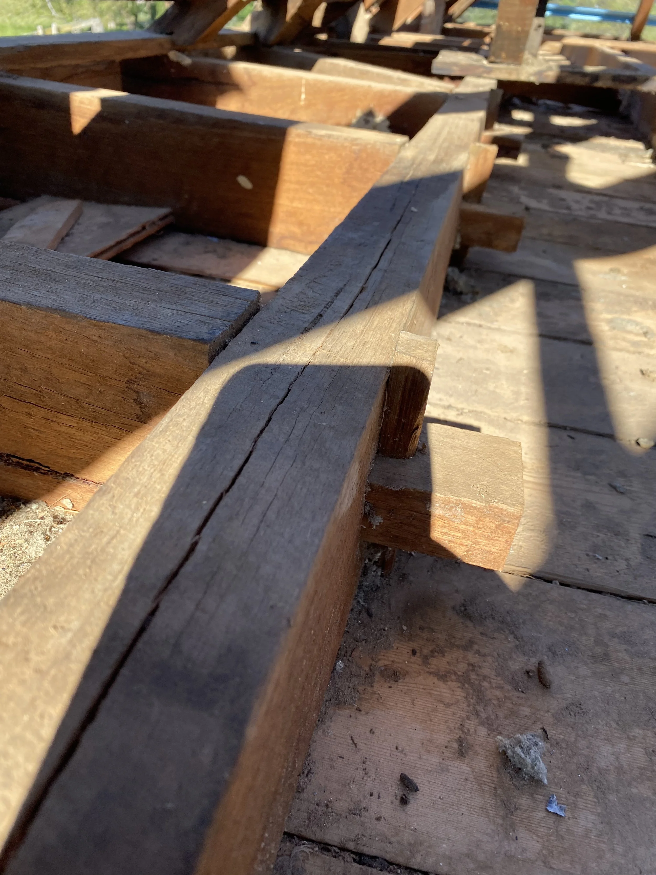 Close-up of a wooden structure, possibly a frame or deck, with sunlight casting shadows on the wood. The wood appears weathered with some dirt and debris on the surface.