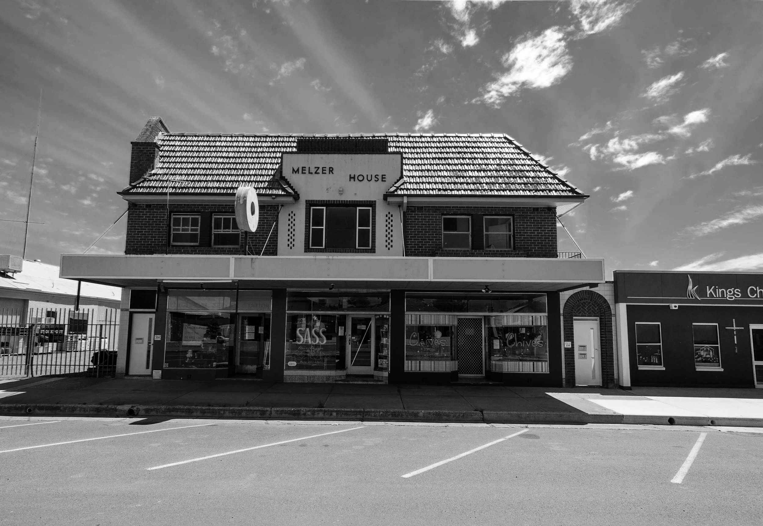 A black and white photo of a two-story brick building with a shingled roof and signage that reads 'Melzer House.' The building has a commercial storefront on the ground floor with large windows displaying signs for businesses named 'SASS,' 'Chives,' and others. An empty parking lot with marked spaces extends in front of the building. The sky above features scattered clouds.