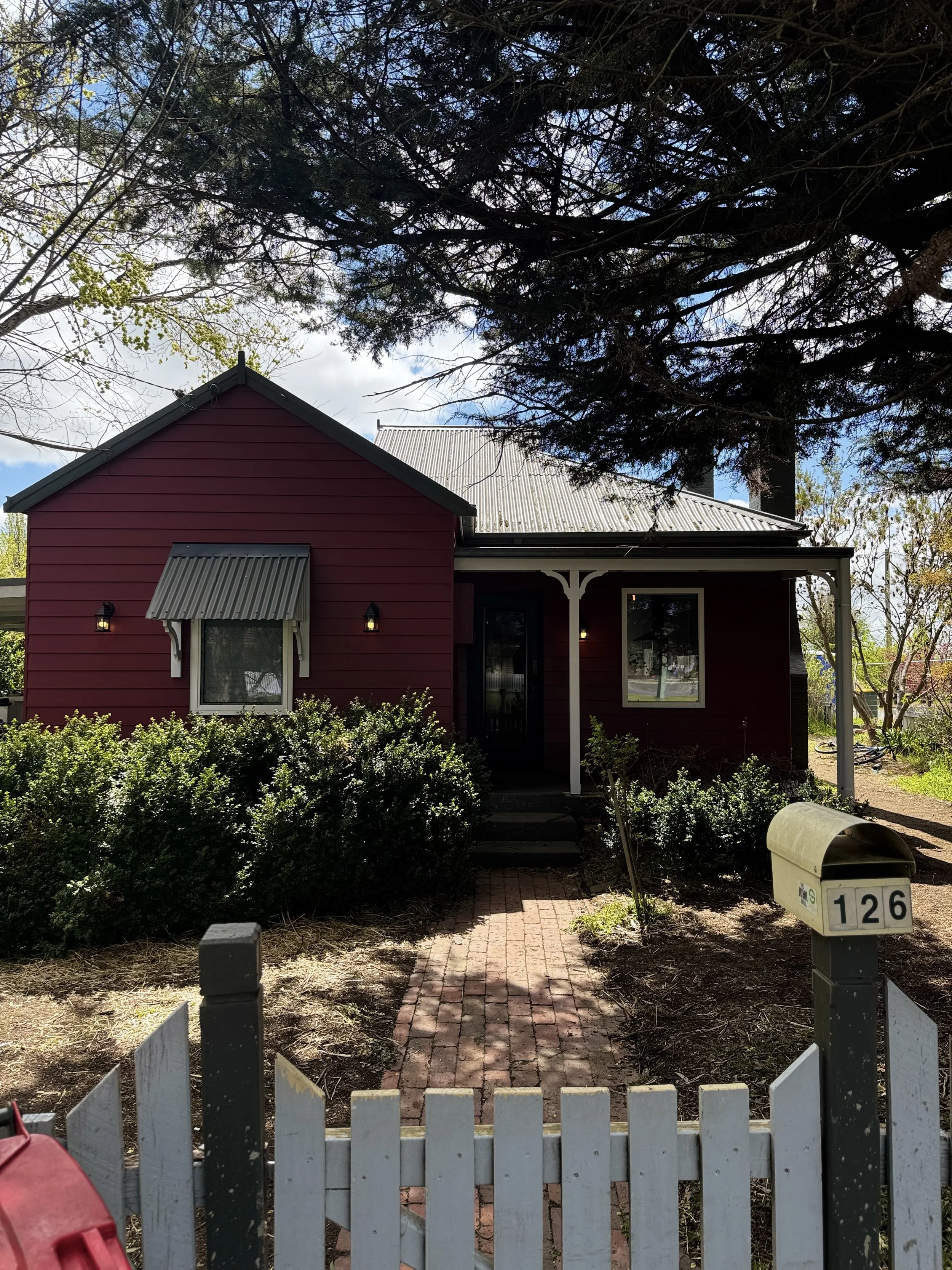 A red house with a white picket fence, a brick pathway leading to the front door, bushes, a mailbox with the number 126, a small porch with white decorative trim, and several trees in the yard.