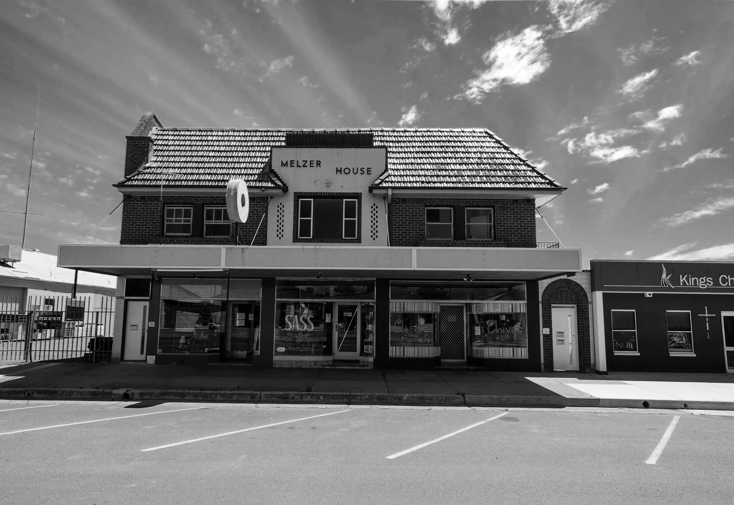 Black and white photo of a two-story brick building with a tiled roof, labeled 'Melzer House', on a street with a parking lot in the foreground.
