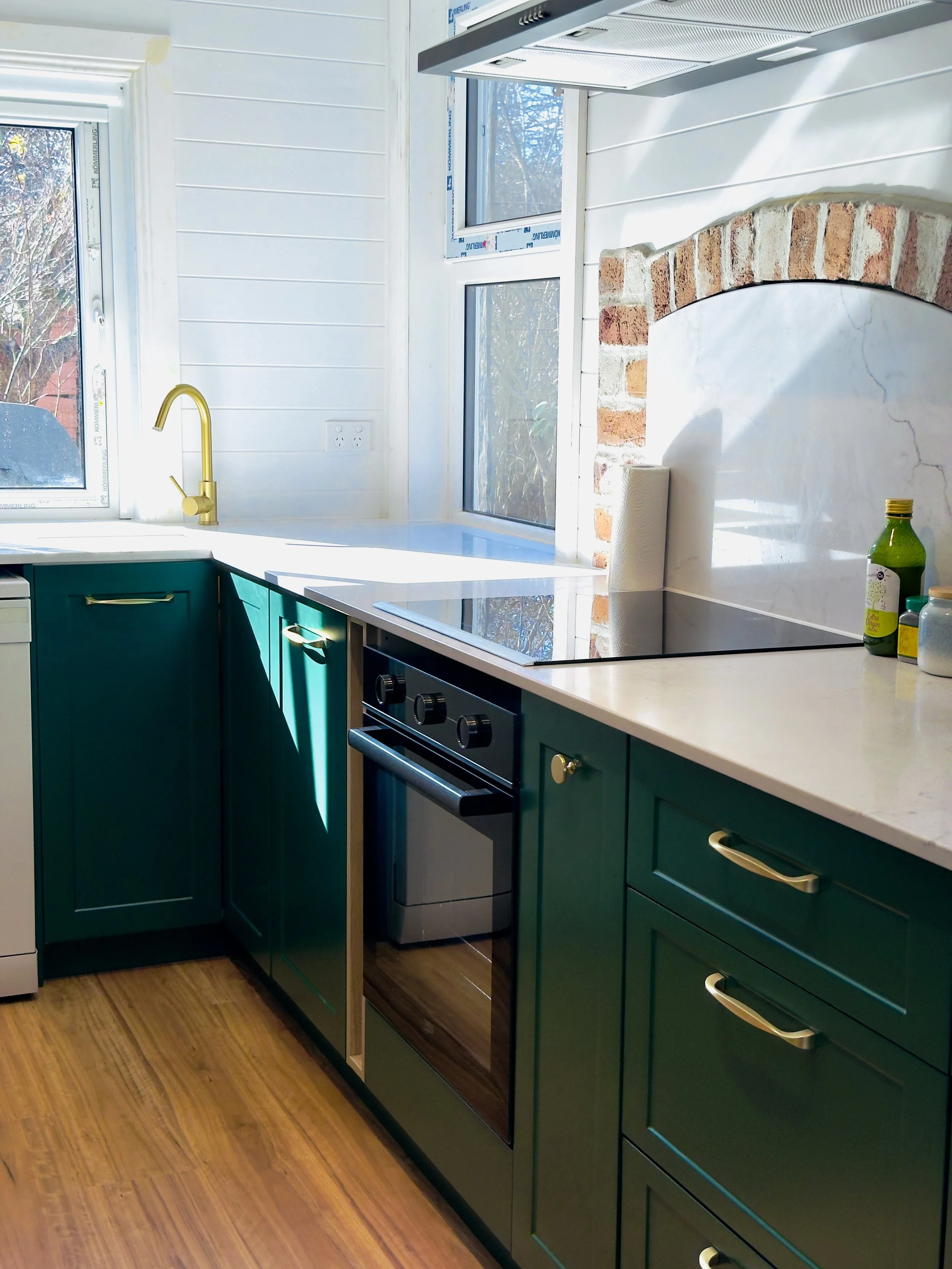 Modern kitchen with green cabinets, white countertop, and brick backsplash. There are windows natural light, a paper towel roll, and a bottle on the counter. An oven is built into the cabinetry.