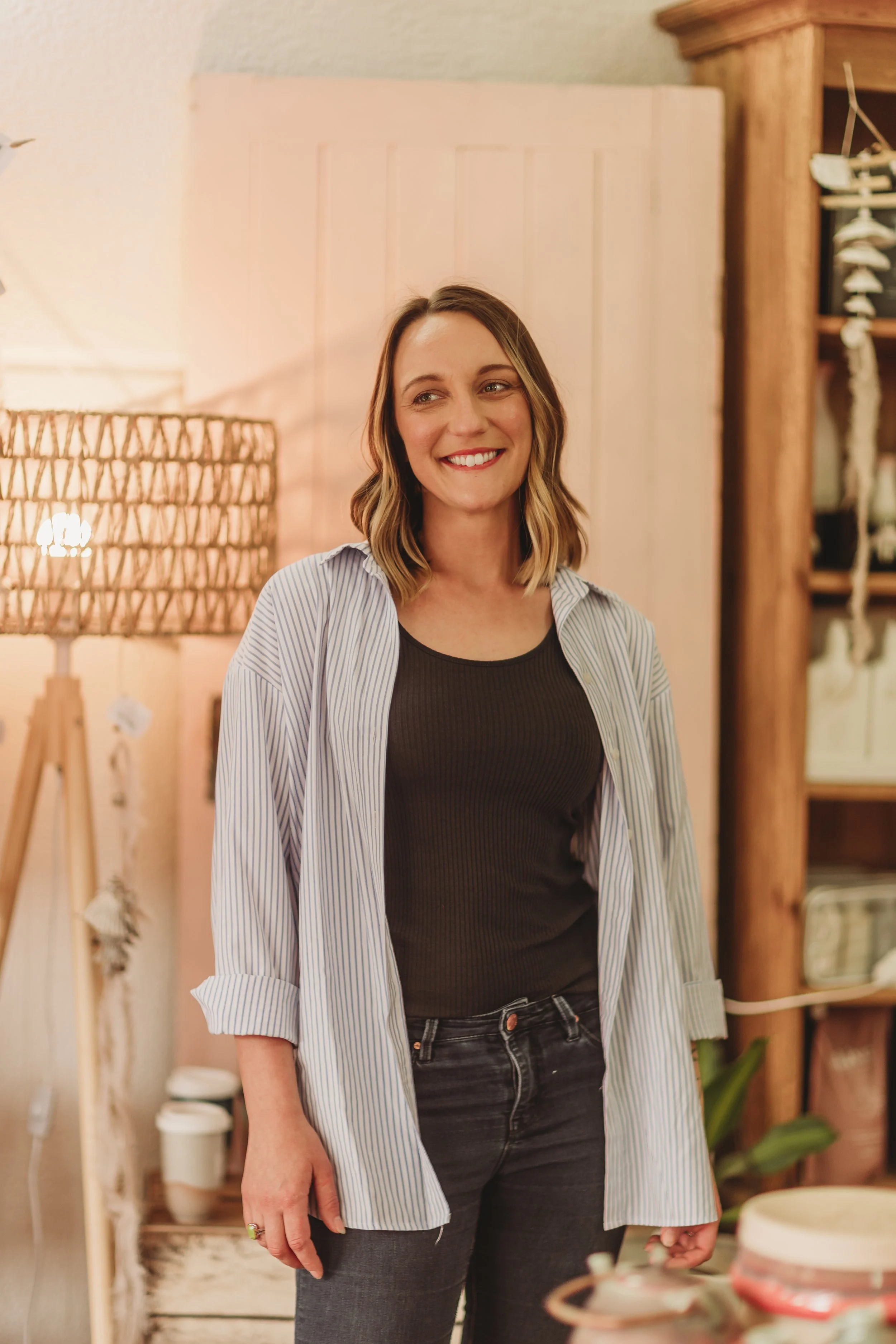 A woman with shoulder-length wavy hair, wearing a black top and a light blue striped button-up shirt, smiling while standing in a cozy home environment with warm lighting, wooden furniture, and decorative items in the background.