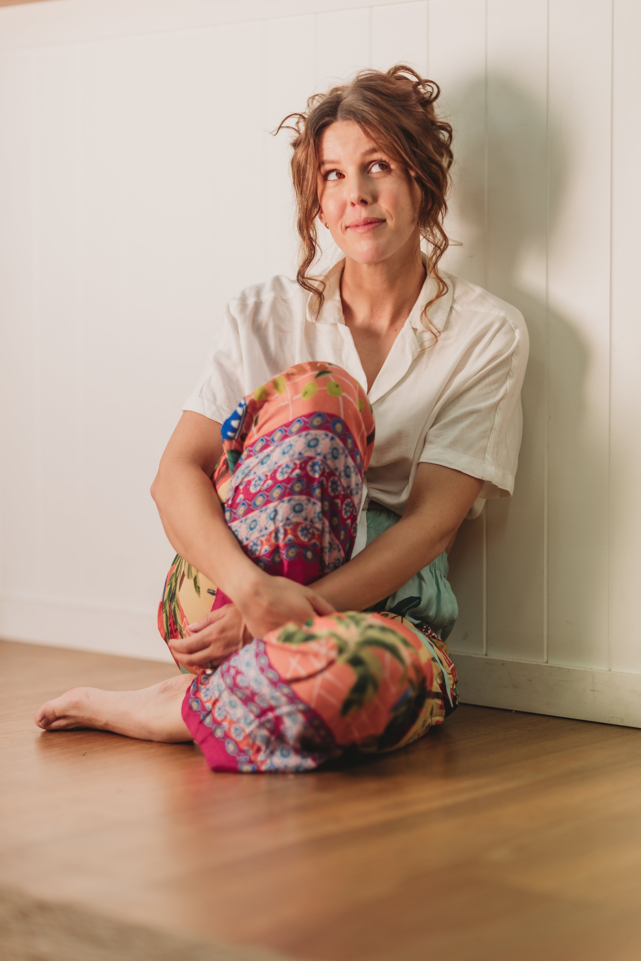 A woman with curly brown hair sitting on a wooden floor, hugging her knees with her arms, wearing a white shirt and colorful patterned pants, looking thoughtfully upward, in front of a white wall.