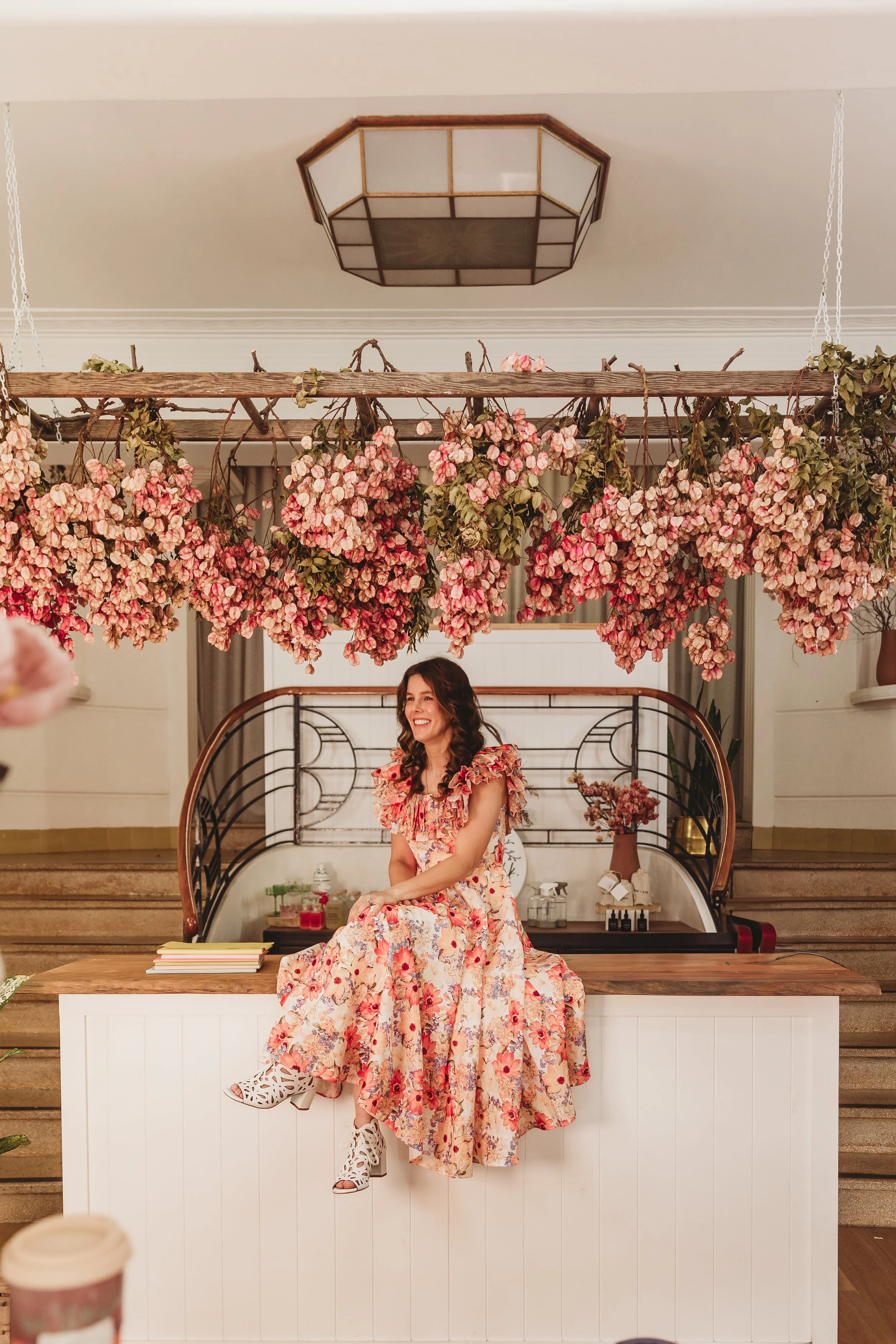 A woman in a floral dress sitting on a counter in a room decorated with hanging pink and green artificial flowers.