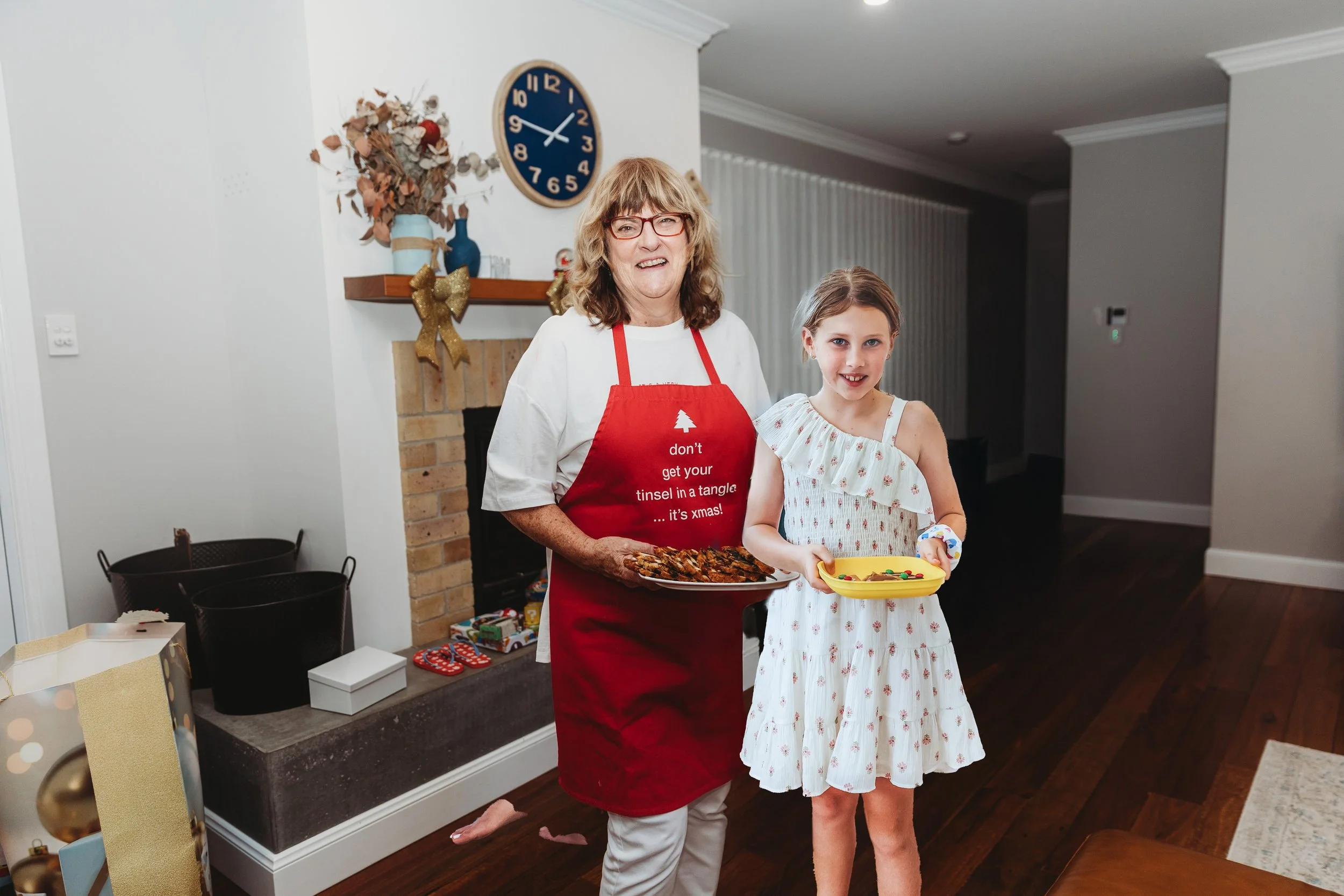 A woman and a young girl at a holiday gathering, the woman wearing a red apron with a Christmas tree and the phrase 'don't get your tinsel in a tangle... it's xmas!'. They are standing near a fireplace, with the woman holding a plate of baked goods and the girl holding a yellow tray with small treats. There are wrapped gifts and decorations in the background, including a wreath and a blue wall clock.