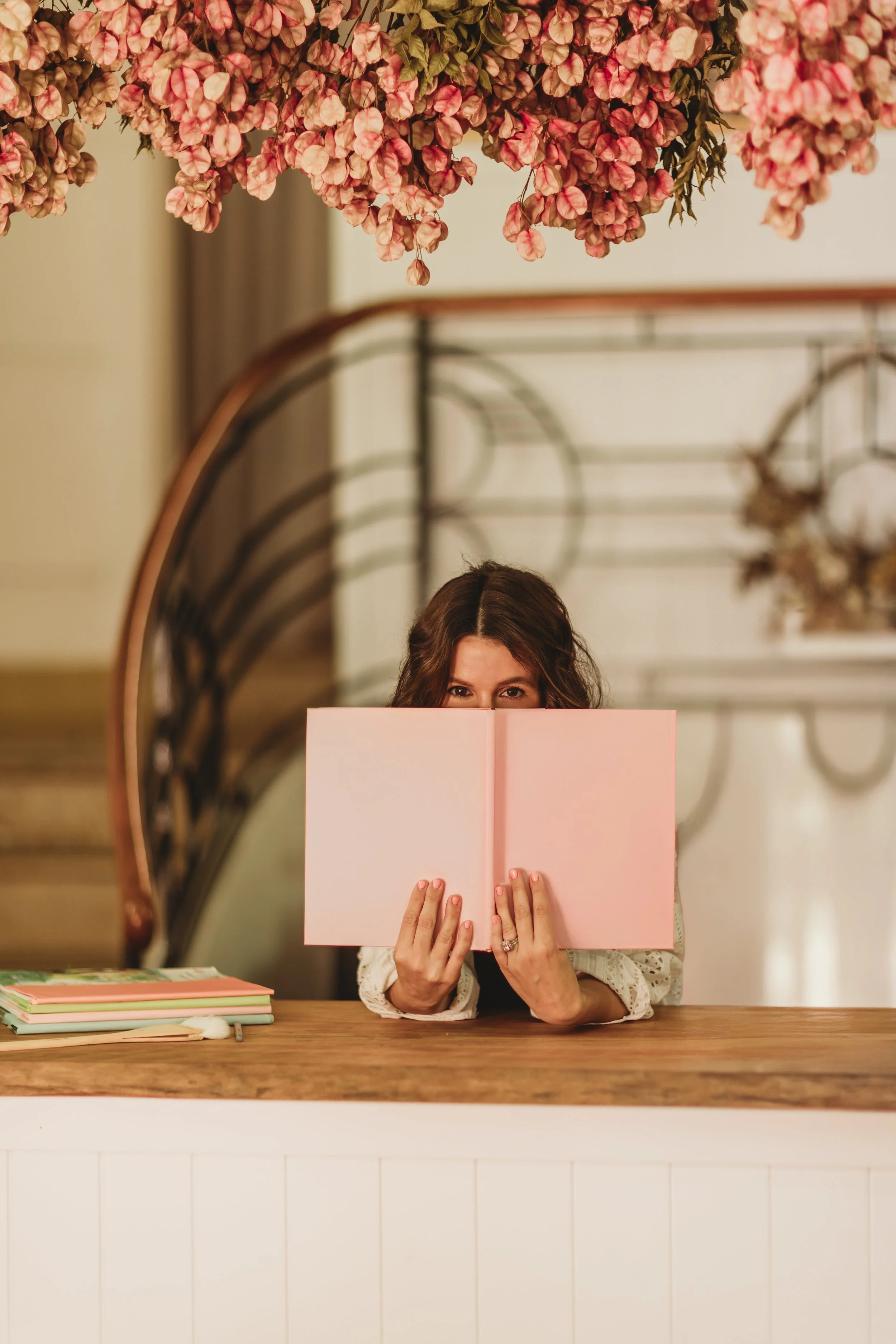 A woman with brown hair holding a pink notebook in front of her face, sitting at a wooden table with more notebooks stacked to her left, in a warmly lit room with a staircase and floral decorations overhead.