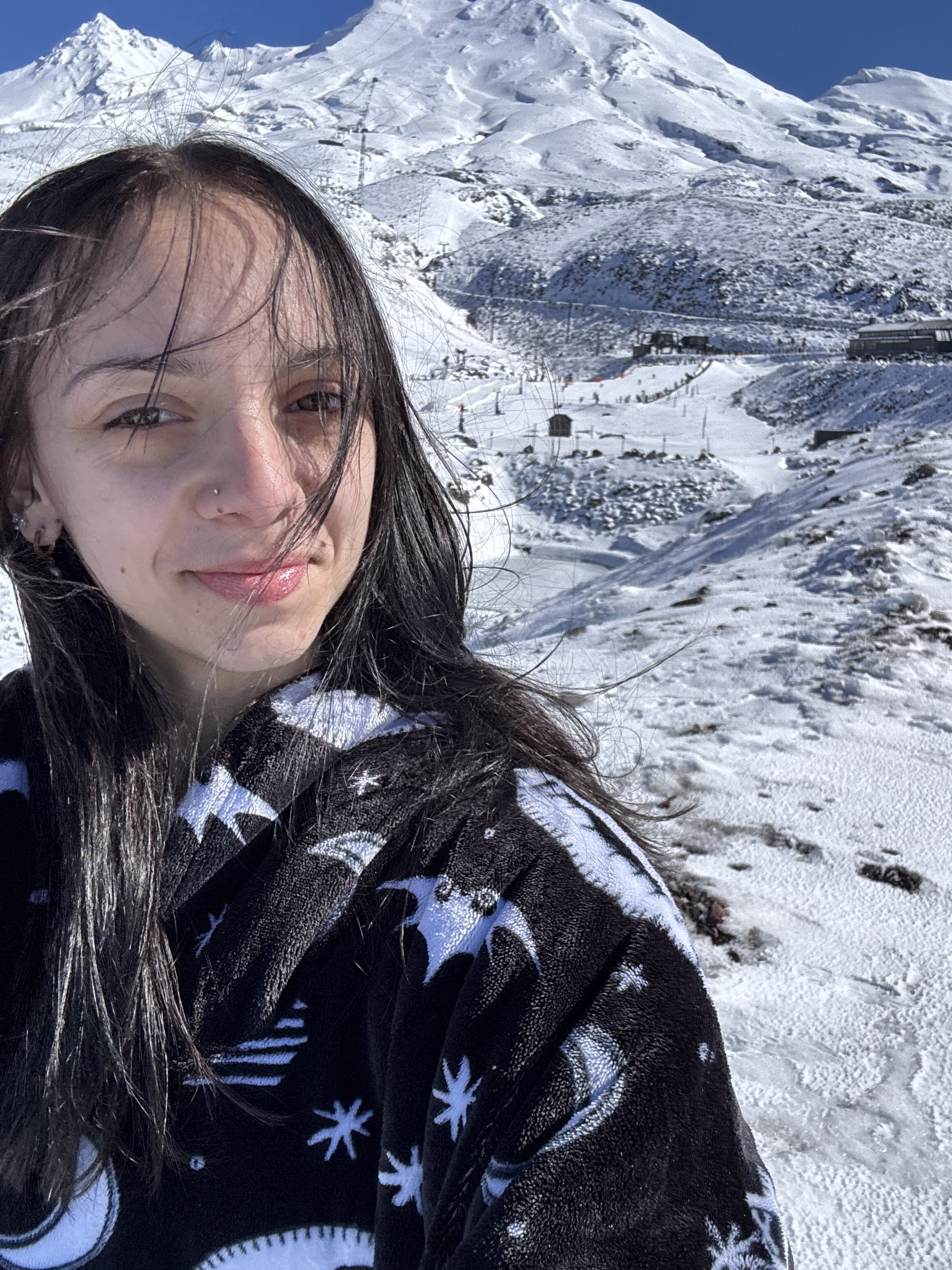 A woman with long dark hair and a nose piercing taking a selfie in a snowy mountainous landscape on a sunny day, wearing a black and white snowflake-patterned sweater.