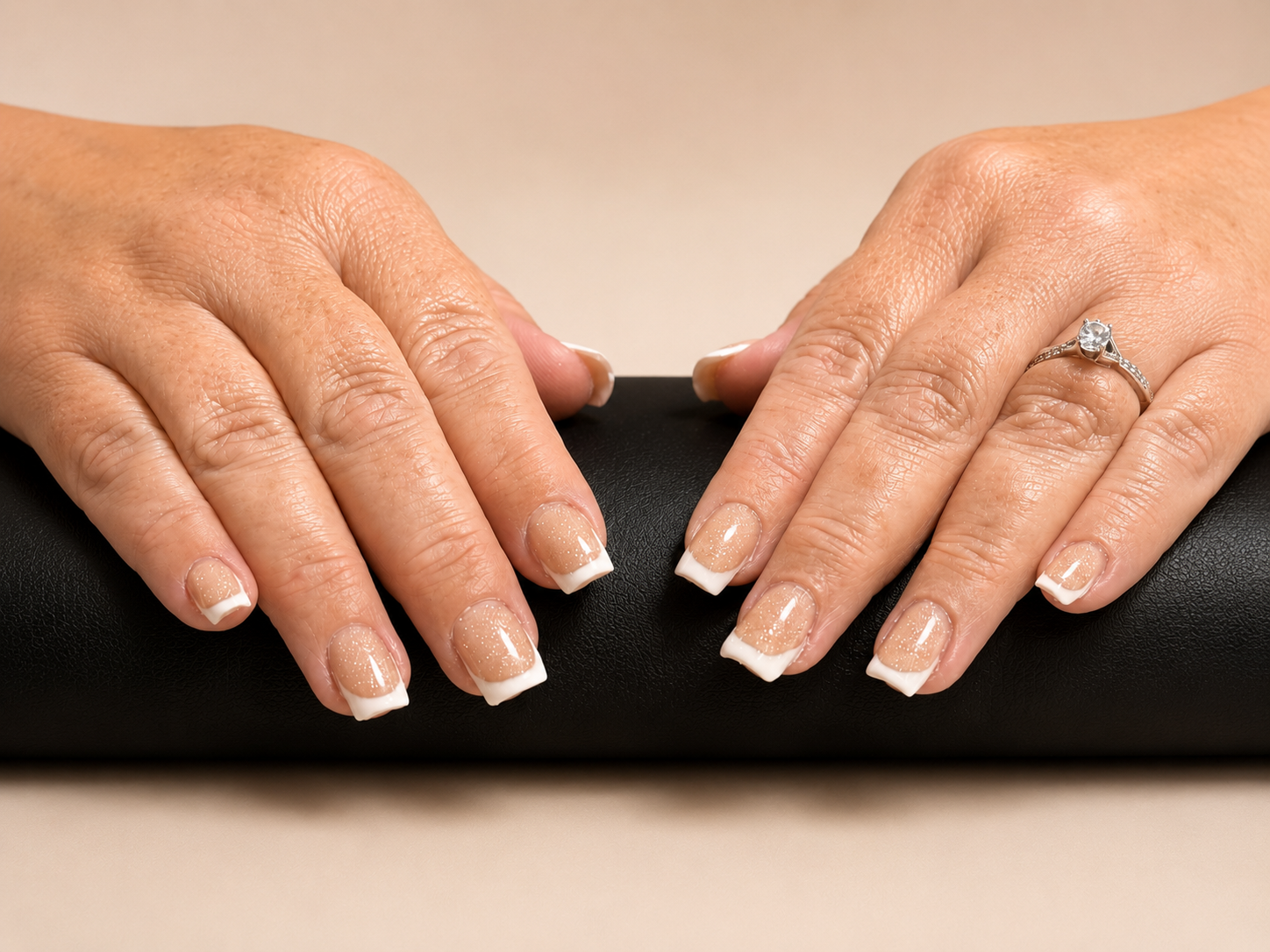 Close-up of a person's hands with manicured nails resting on a black surface. The nails have French tips with glittery beige polish, and the person wears a silver ring with a large stone on the right hand.