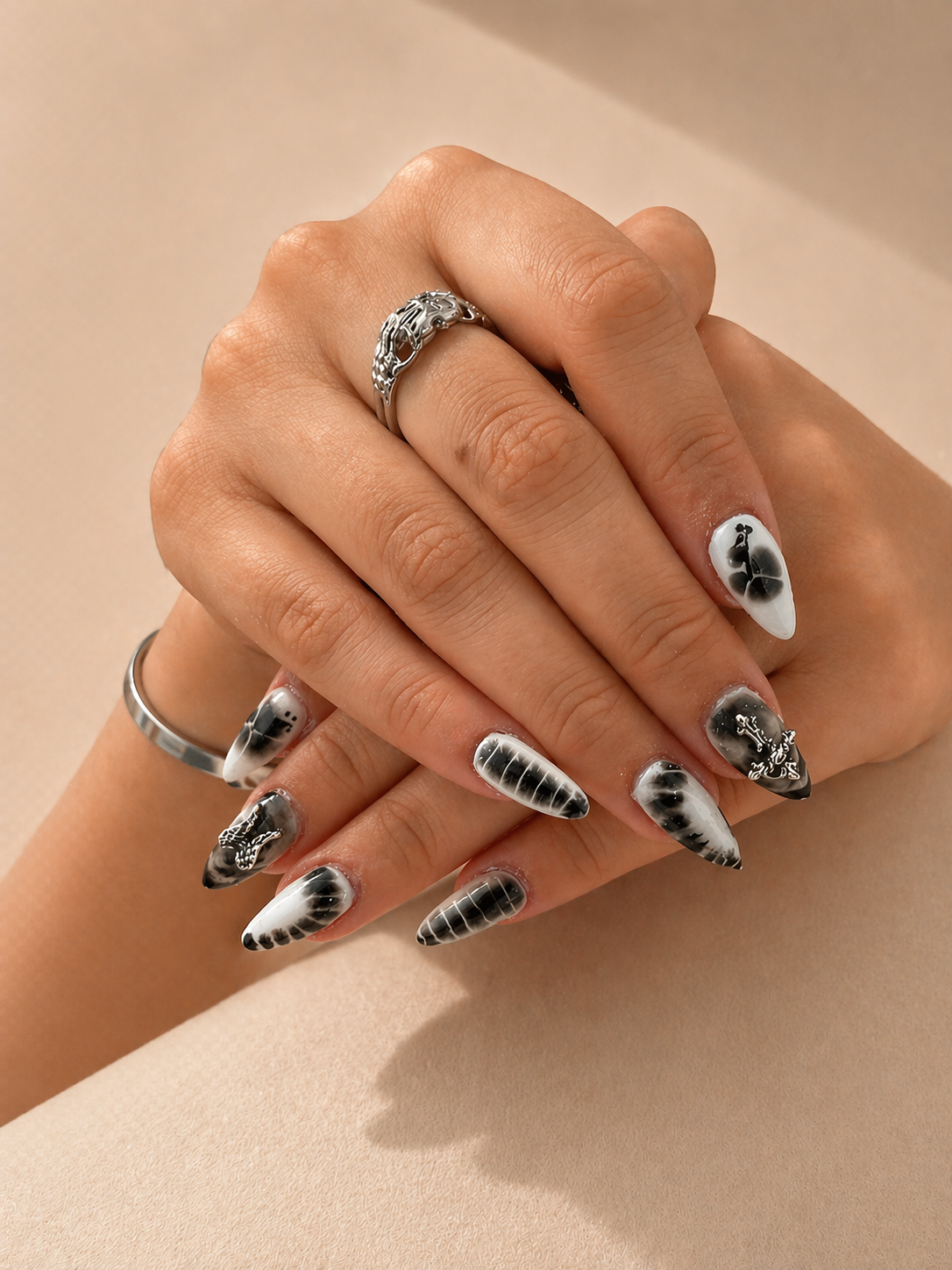 Close-up of a person's hands with black and white marble nail art, wearing silver rings with intricate designs, posed against a beige background.