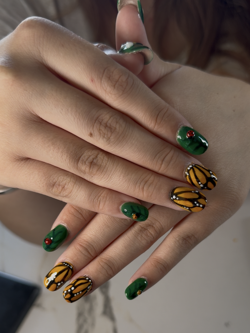 Close-up of hands with intricately painted nail art featuring butterfly and ladybug designs in yellow, black, white, and red on green backgrounds.