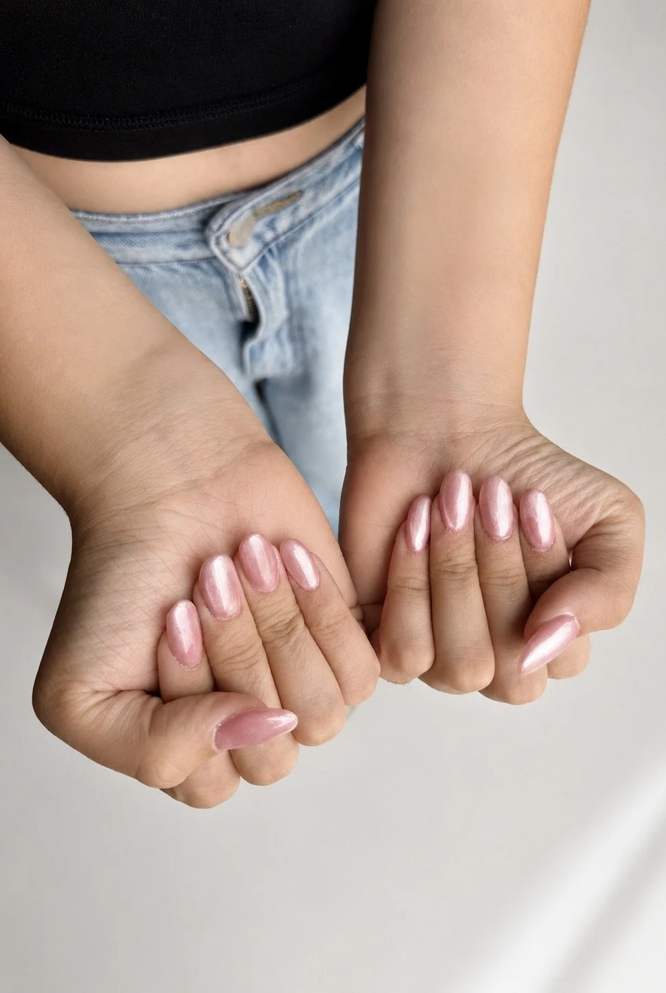 Close-up of a person with pink metallic nails holding their fists together, wearing a black top and light blue jeans.